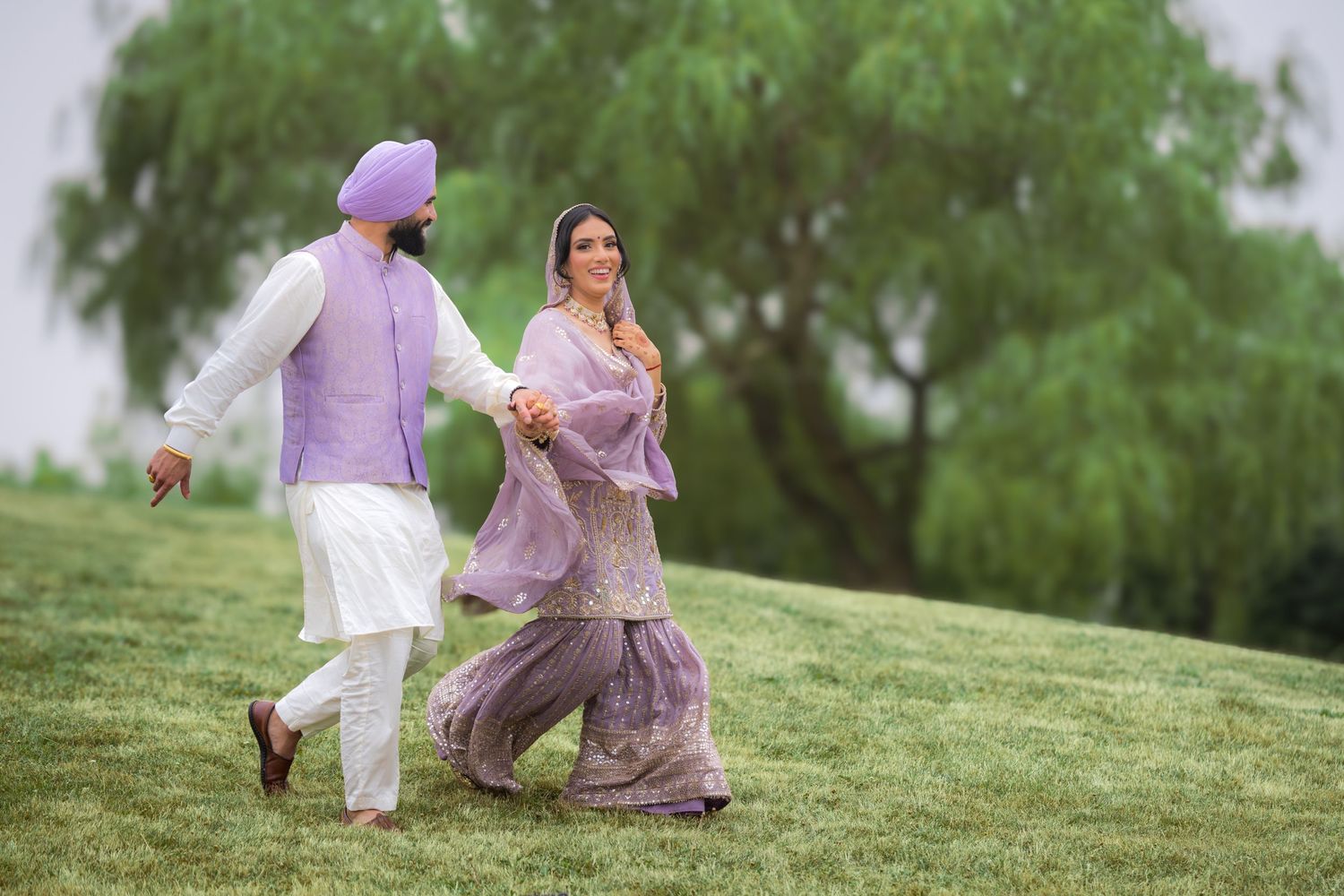 Romantic couple in purple and white traditional attire runs playfully on a grassy slope.