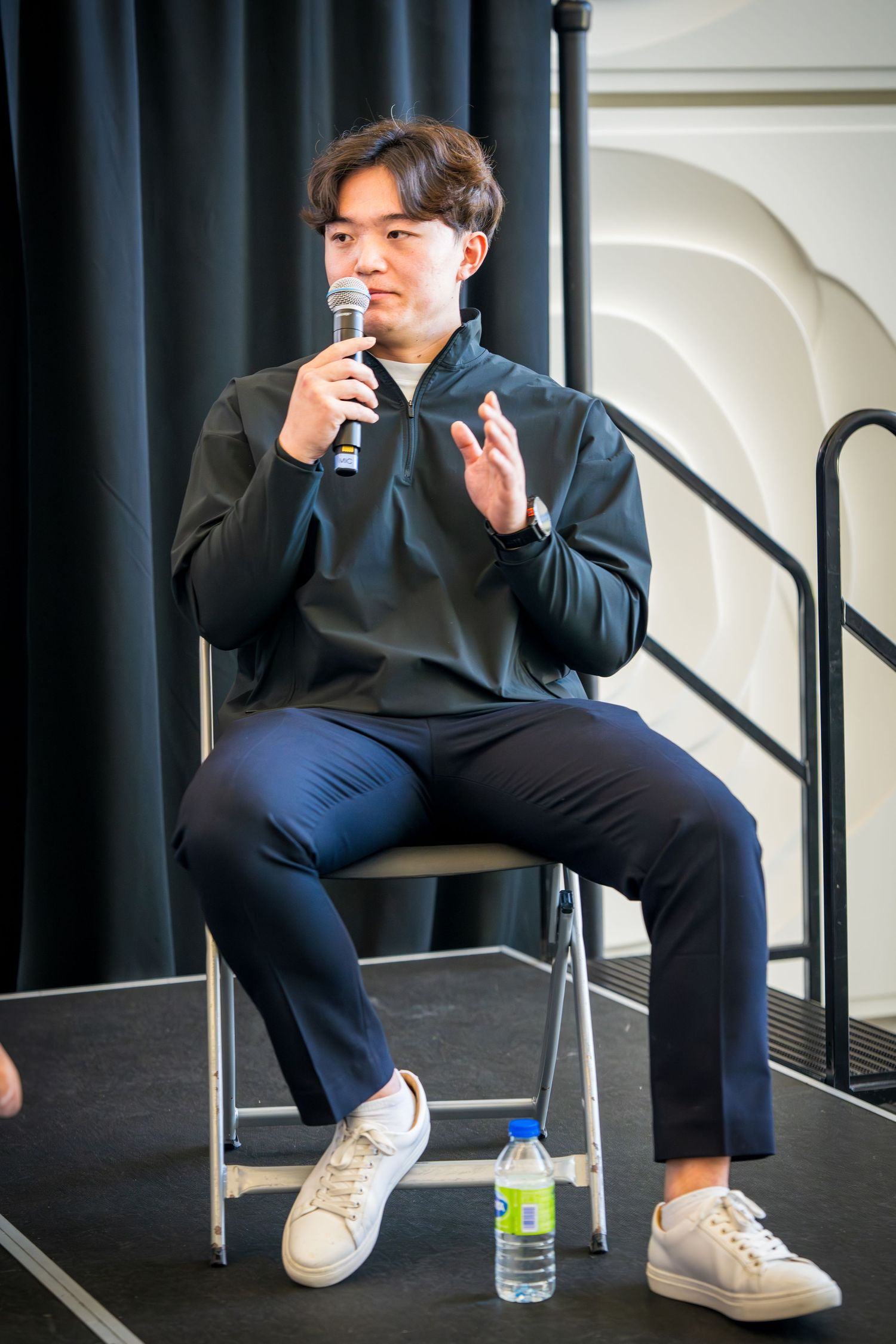 Person in dark clothing seated on chair taking a drink from water bottle during event.