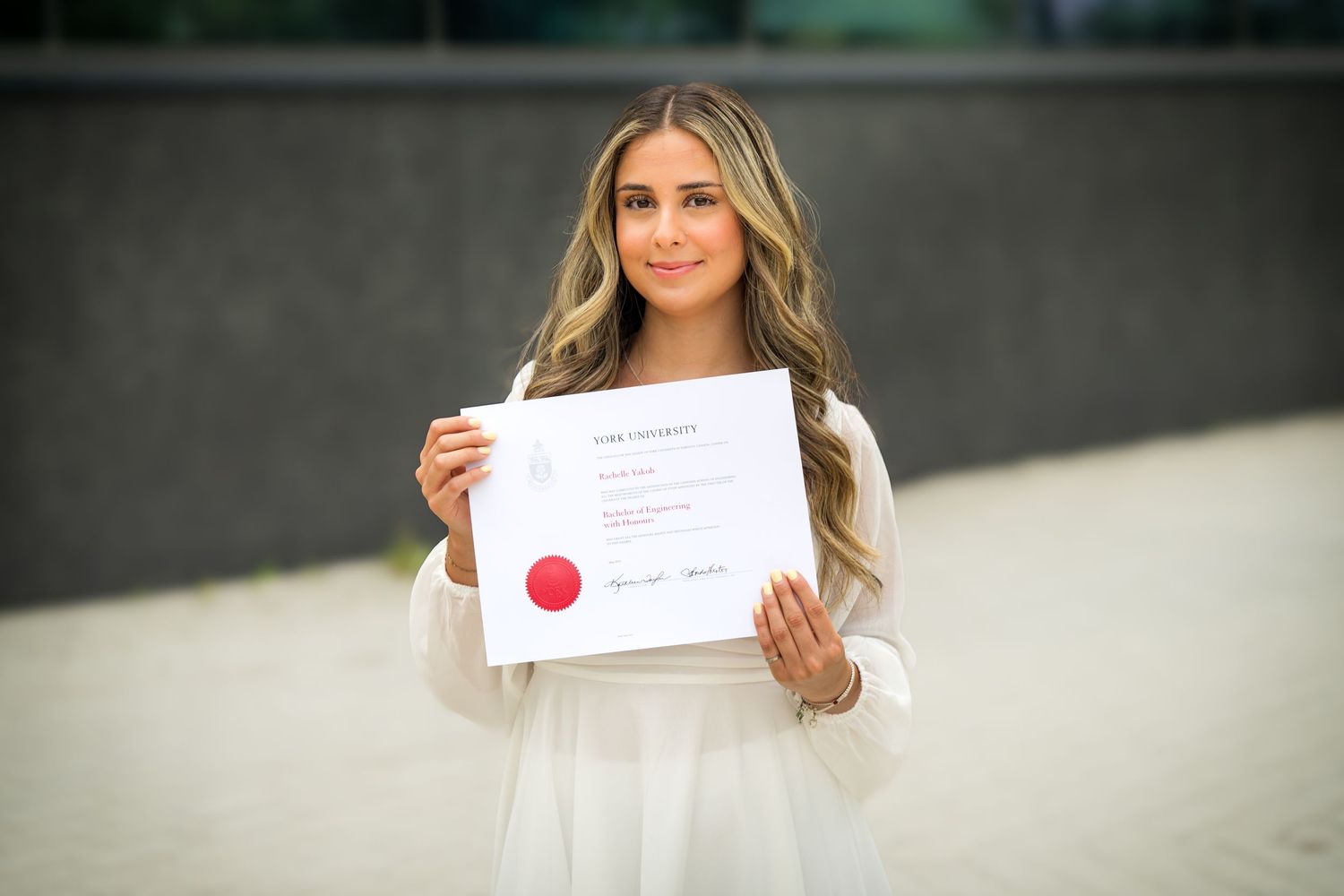 Graduate in flowing white dress proudly displays university diploma with red seal.