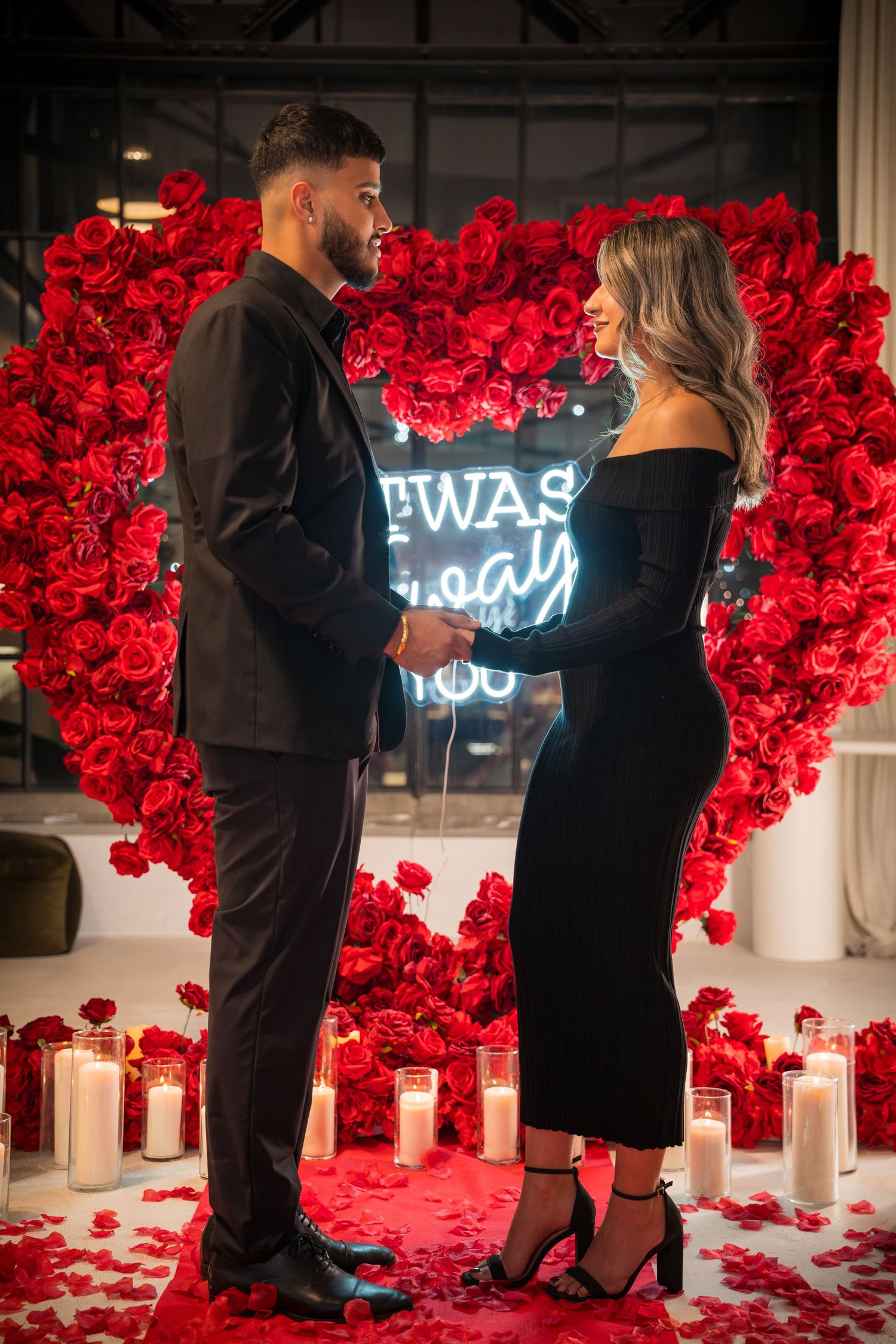 Romantic proposal setup with red roses and neon sign against white walls and scattered petals on floor.