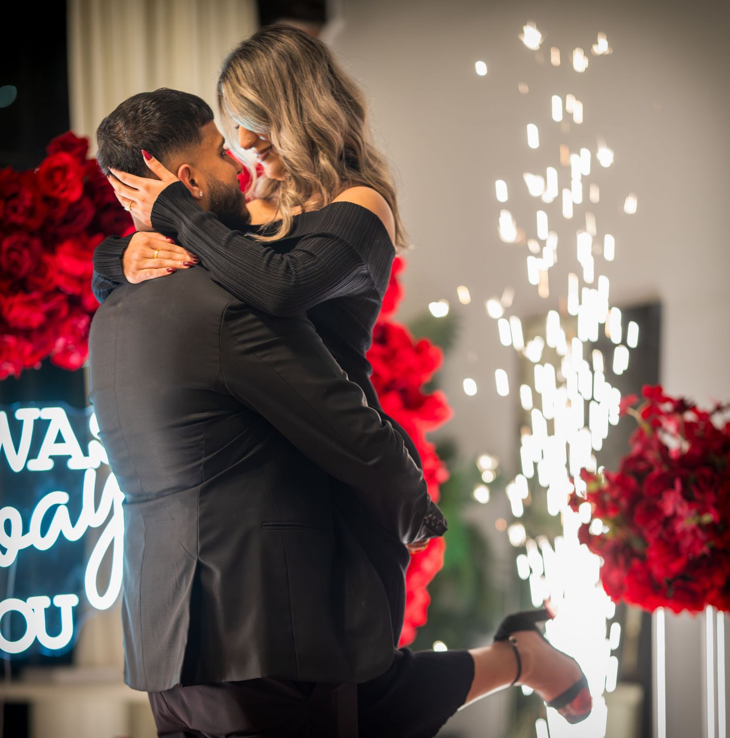 Romantic proposal moment with sparklers and red rose decorations in the background.