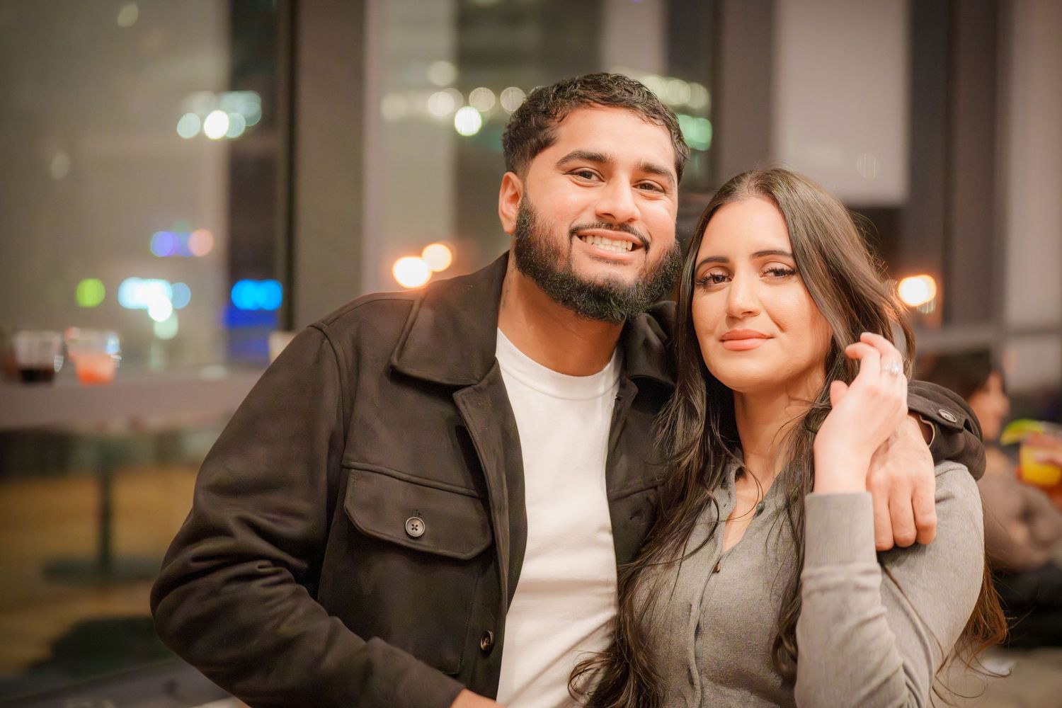 Cozy indoor portrait of a couple smiling with city lights visible in the background.