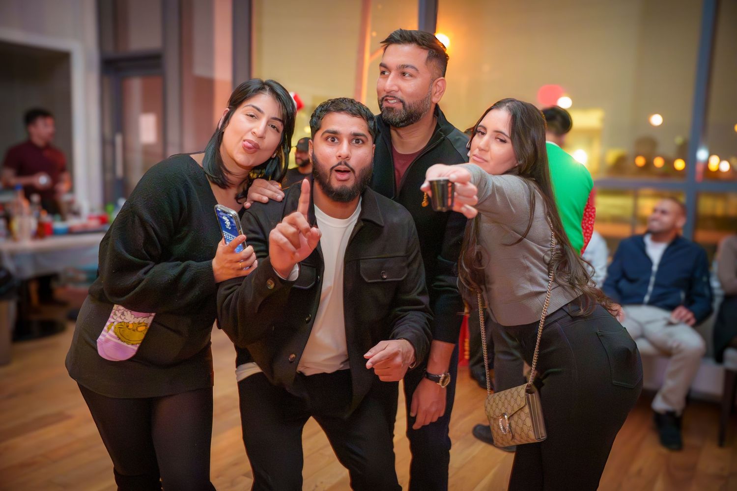 Group of friends posing playfully with drinks at an indoor party with warm lighting.