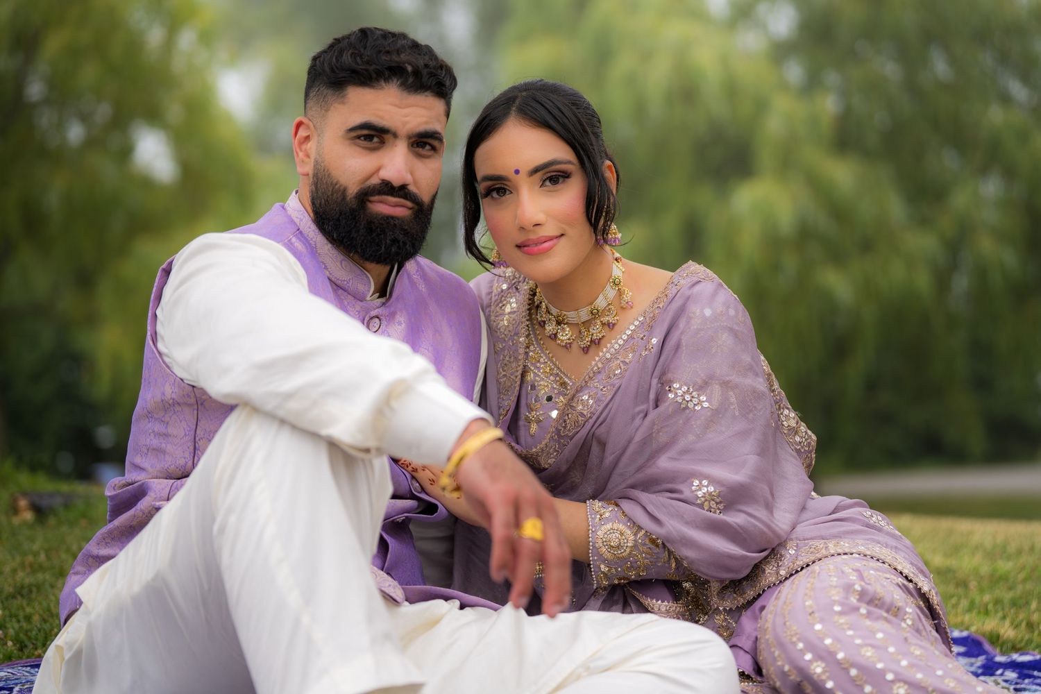 Couple in traditional purple and white South Asian attire posing together outdoors in a garden setting.