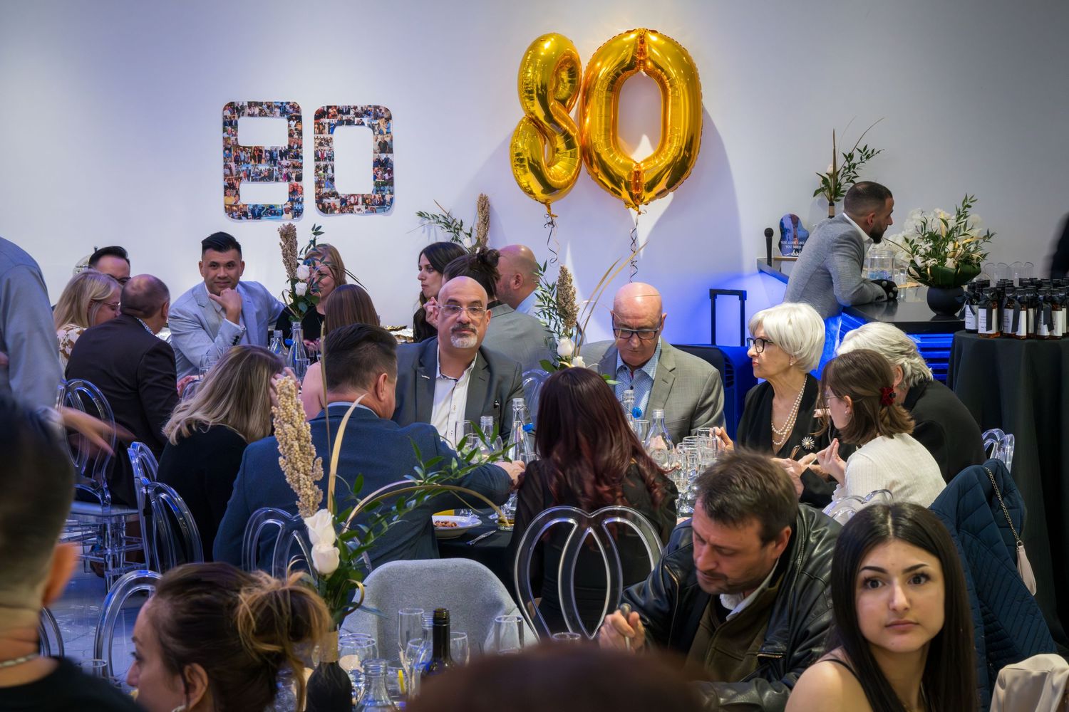 Festive gathering with gold 80th birthday balloons and guests seated at decorated tables.