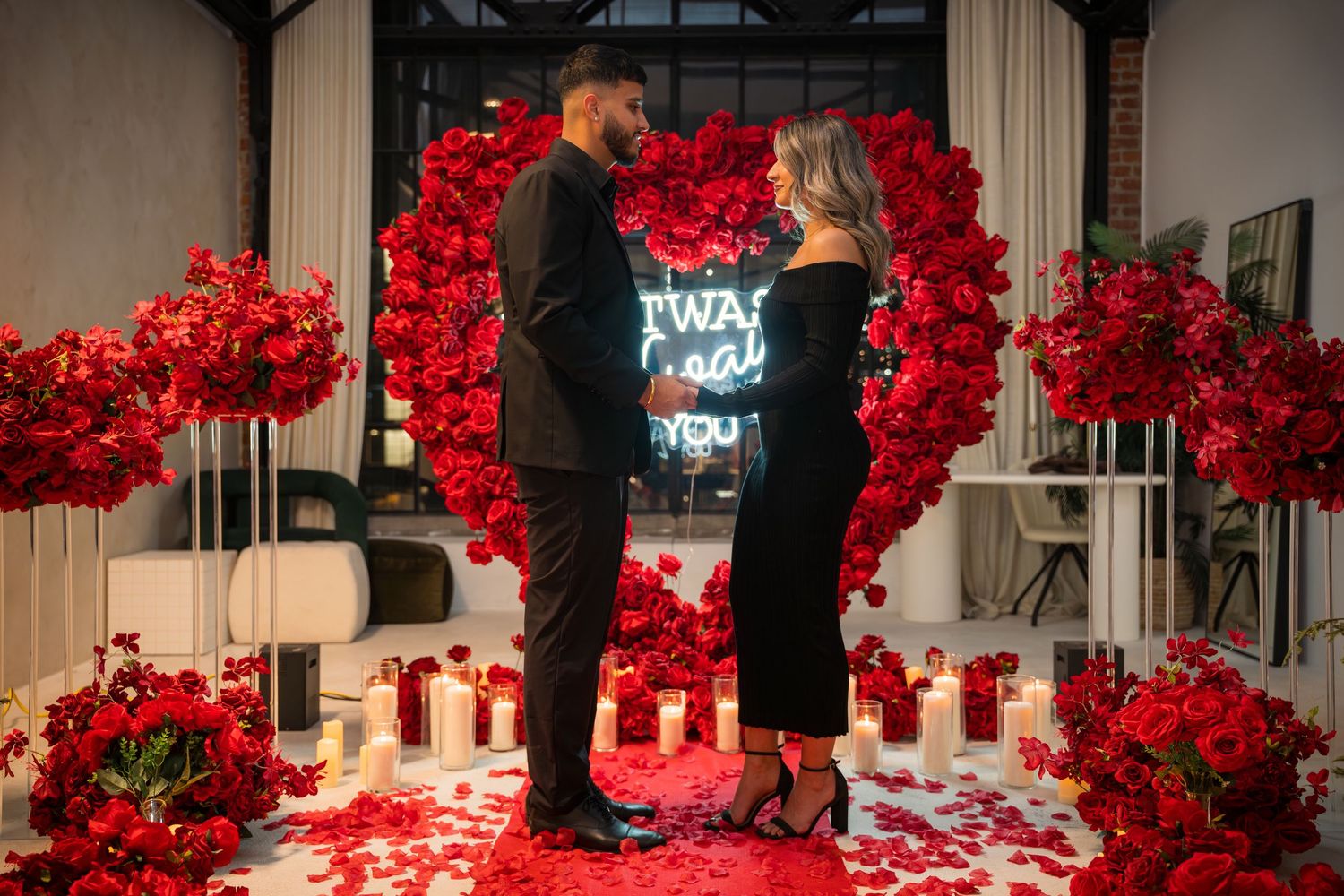 Wide shot of romantic proposal setup with red rose walls and candle-lined pathway.