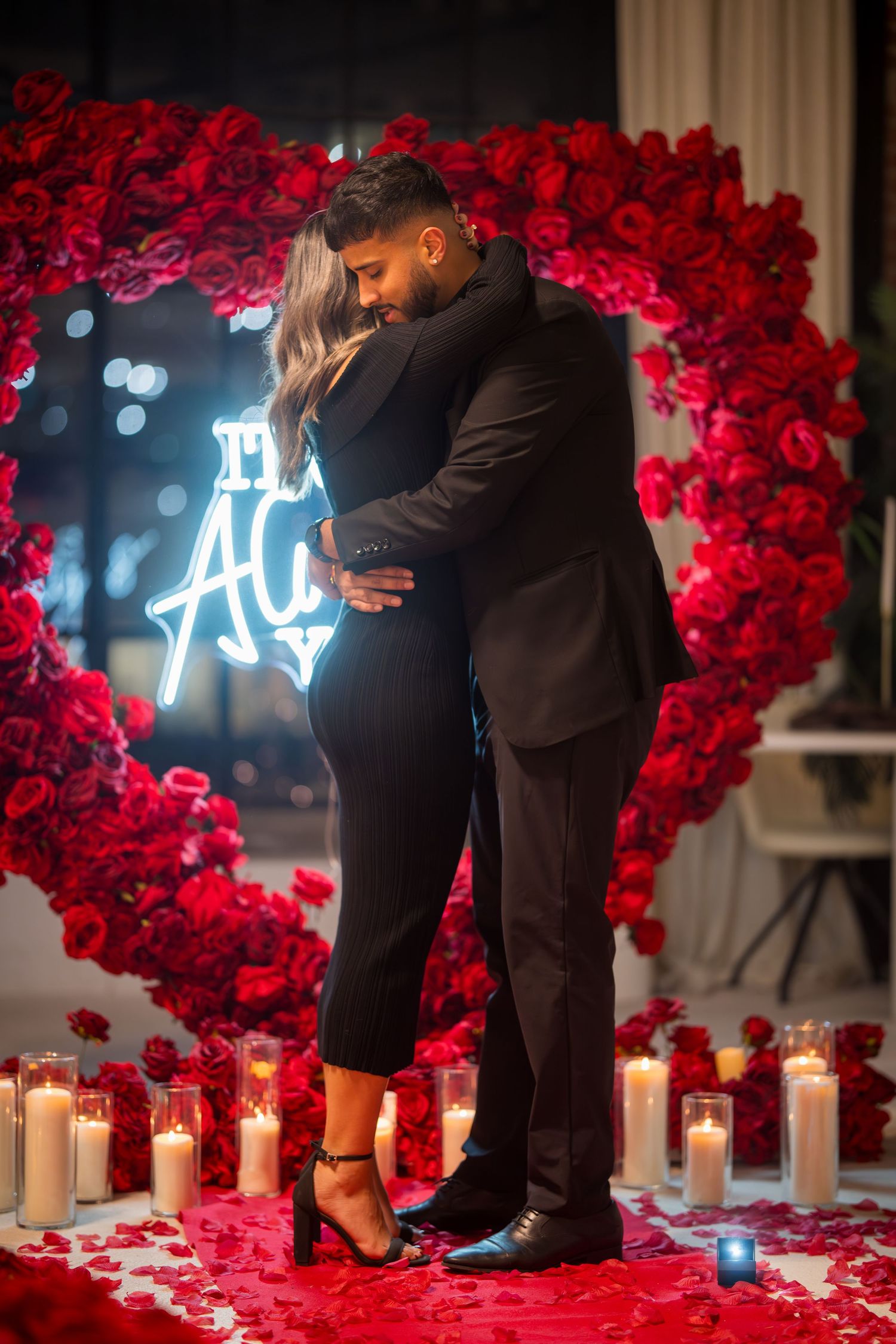 Romantic proposal scene with red rose wall, candles, and illuminated neon sign.