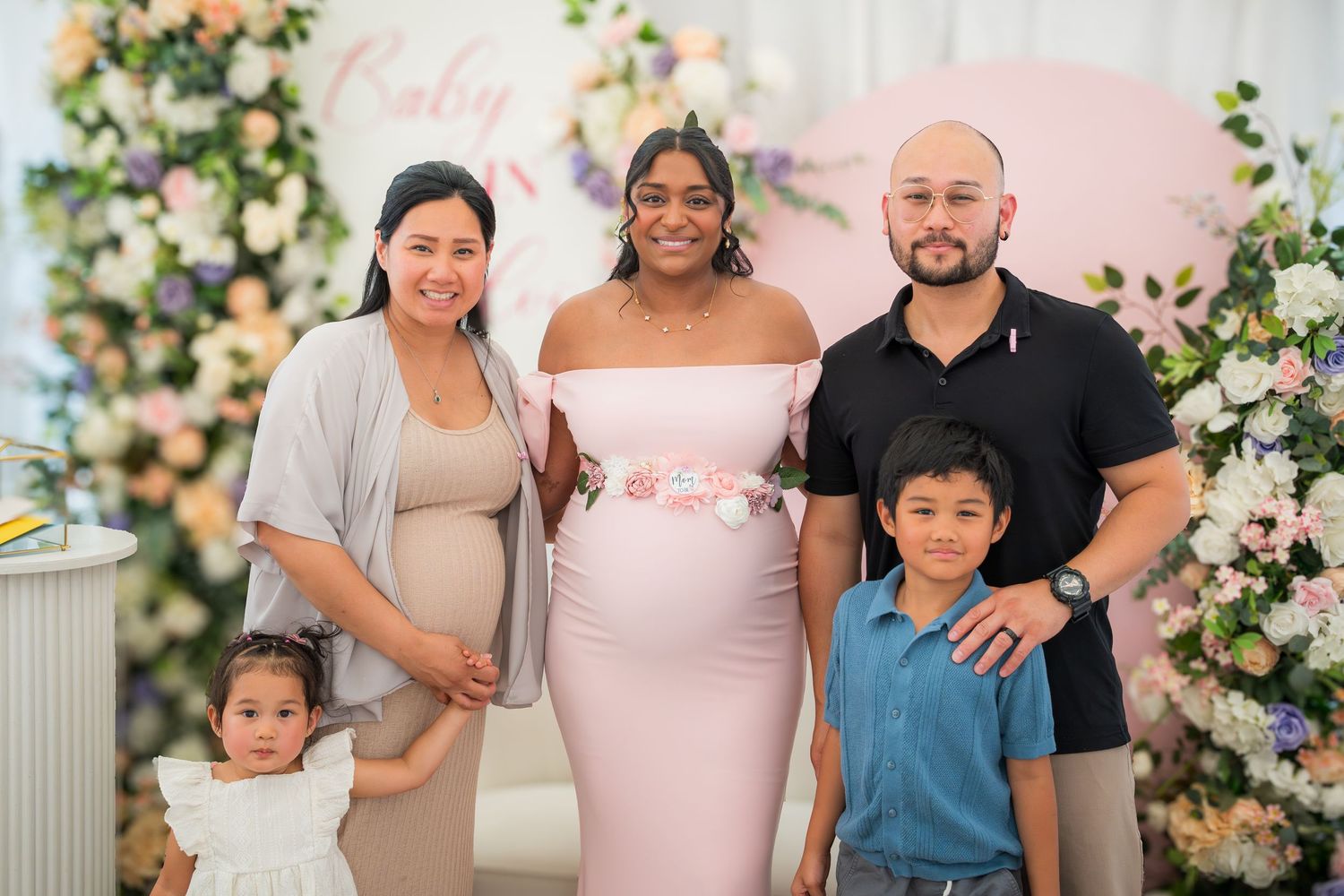 Family photo at a baby shower with floral arch and pink backdrop.