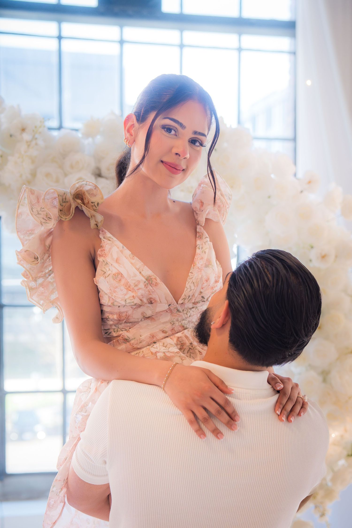 Woman in floral dress being lifted by man in front of white flower heart backdrop.