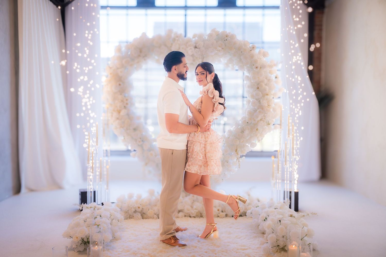 Happy engaged couple dancing together in front of a glowing white floral heart arch with fairy lights indoors.