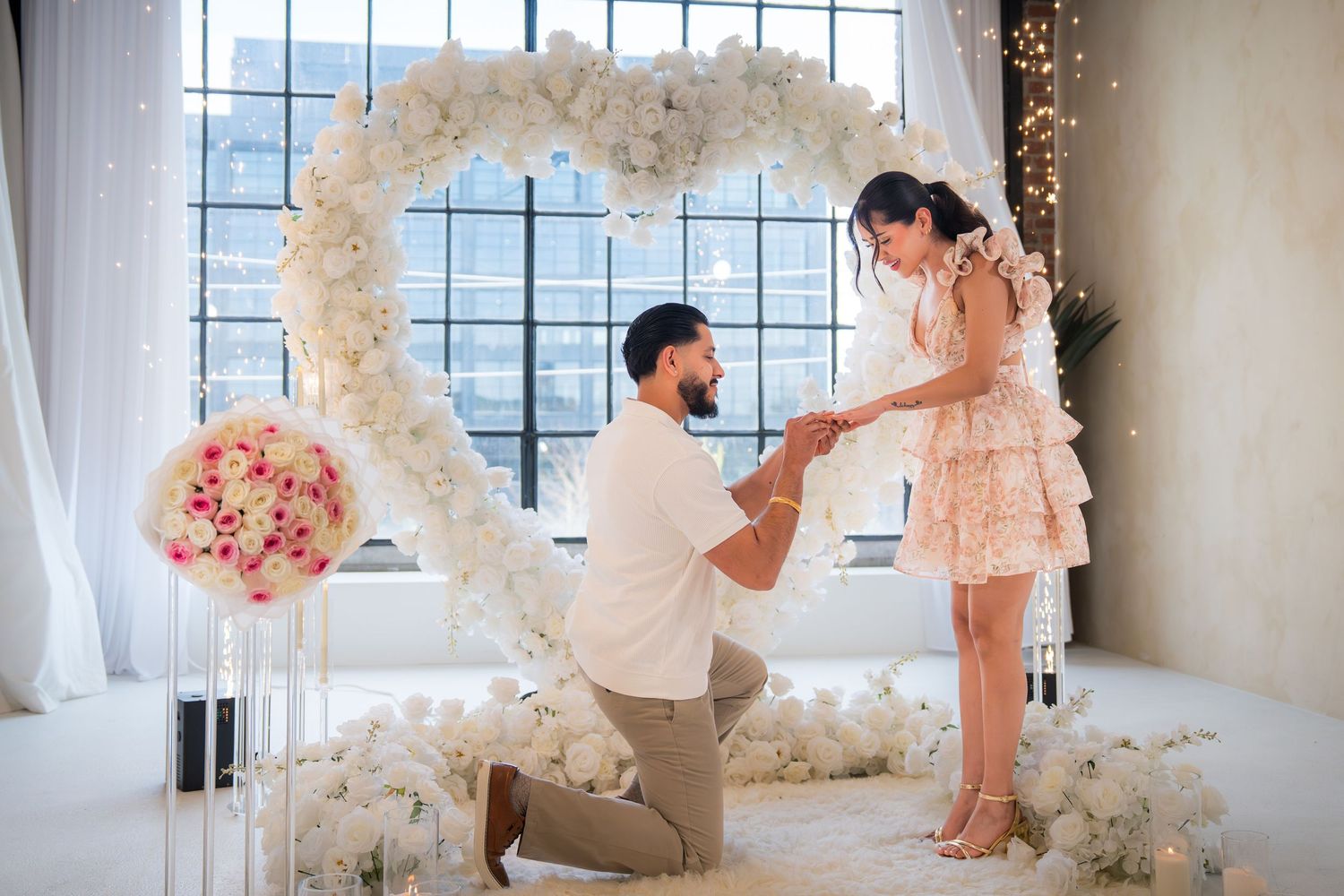 Romantic marriage proposal with man kneeling before woman beneath white flower heart arch indoors.