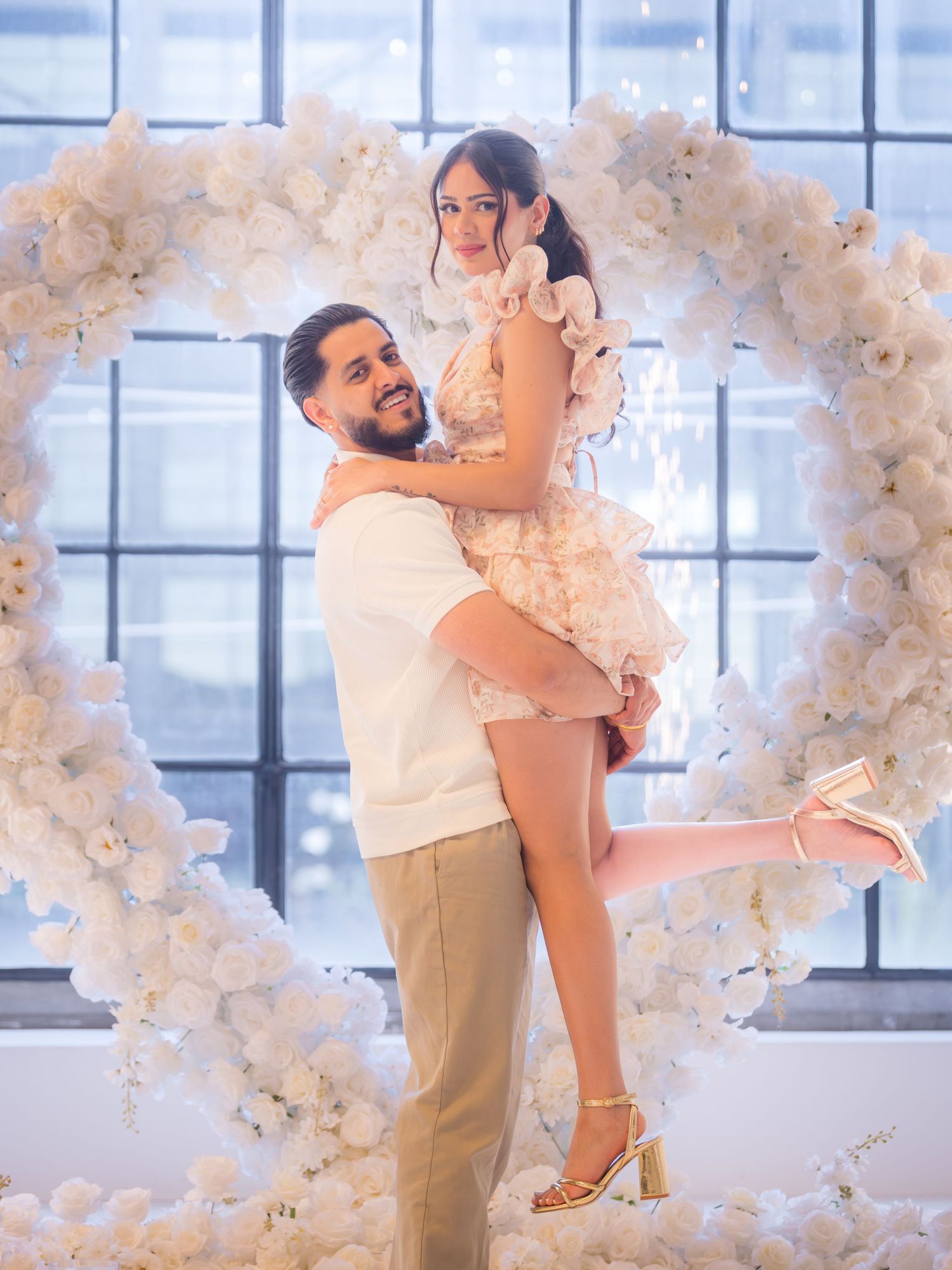 Joyful couple embracing in front of large white floral heart arch at engagement celebration.