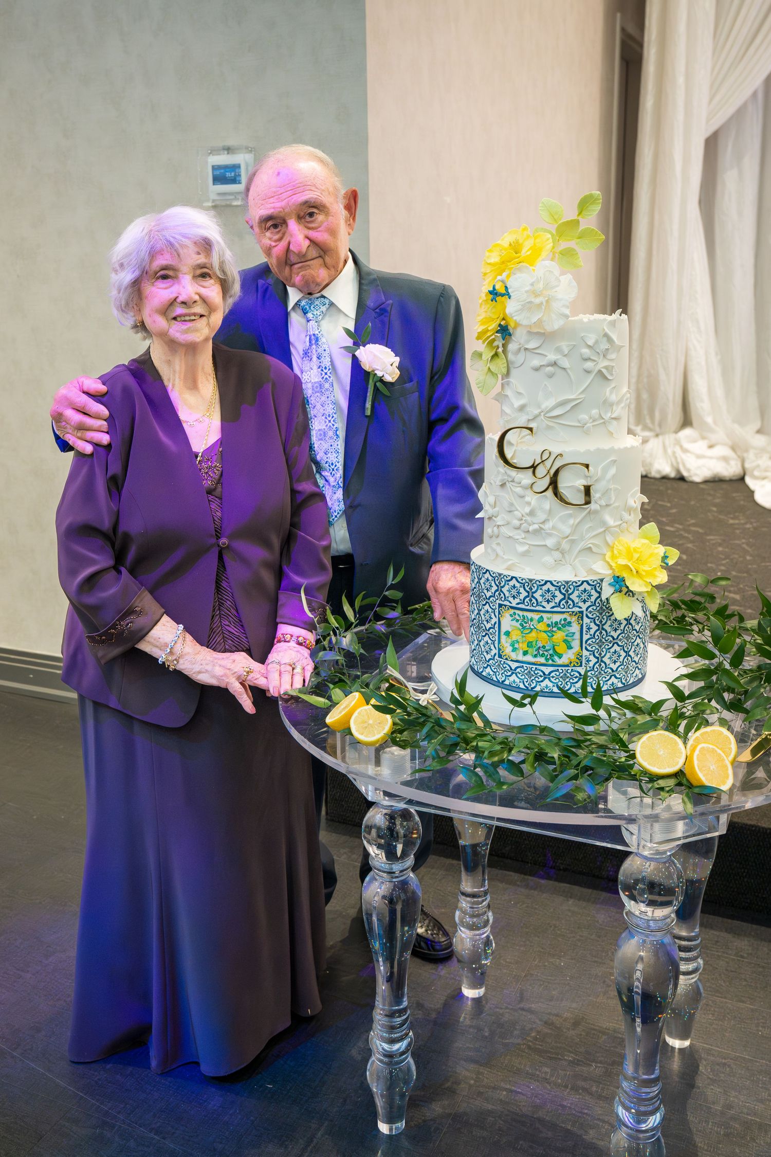 Elderly couple in purple and blue formal attire posing beside a three-tier wedding cake decorated with lemons.