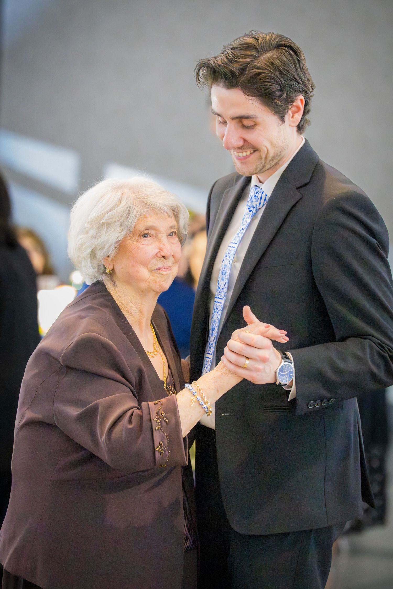 Elderly woman in brown jacket holds hands with young man in dark suit at formal event.