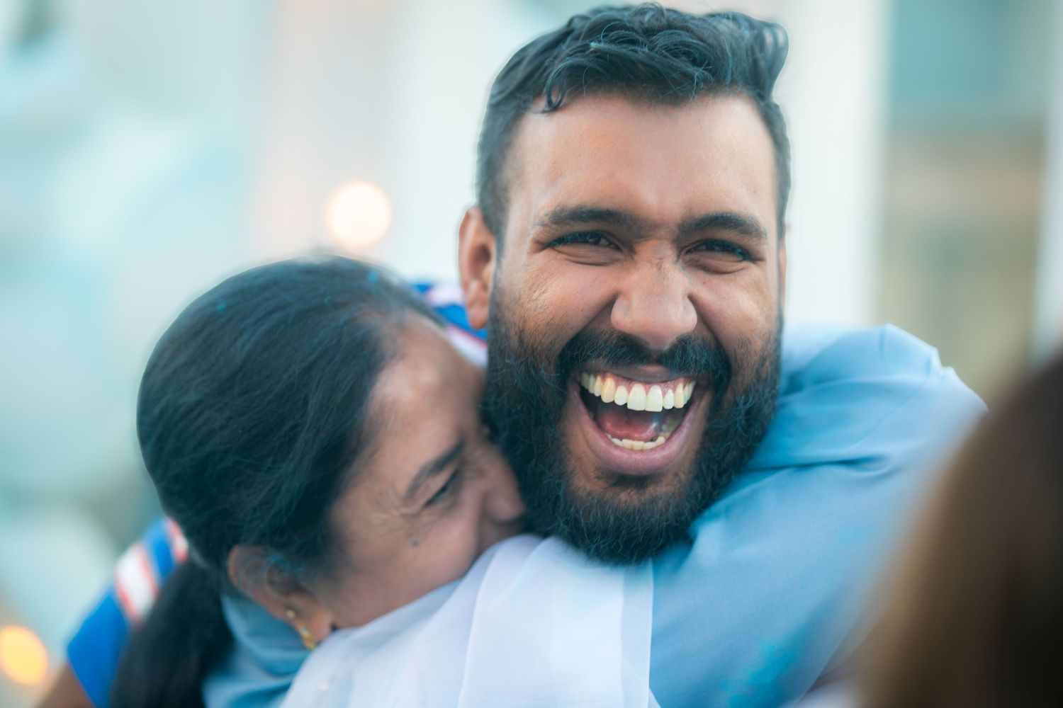 A joyful man with a beard laughs while a woman hugs him warmly, both wearing light blue clothing outdoors.