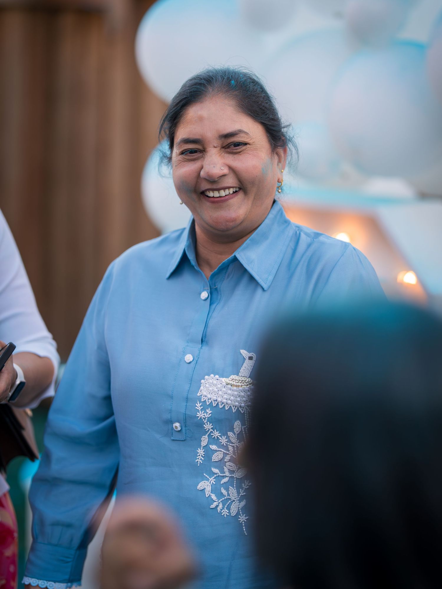 A smiling middle-aged woman in a blue embroidered shirt at a balloon-decorated outdoor event.