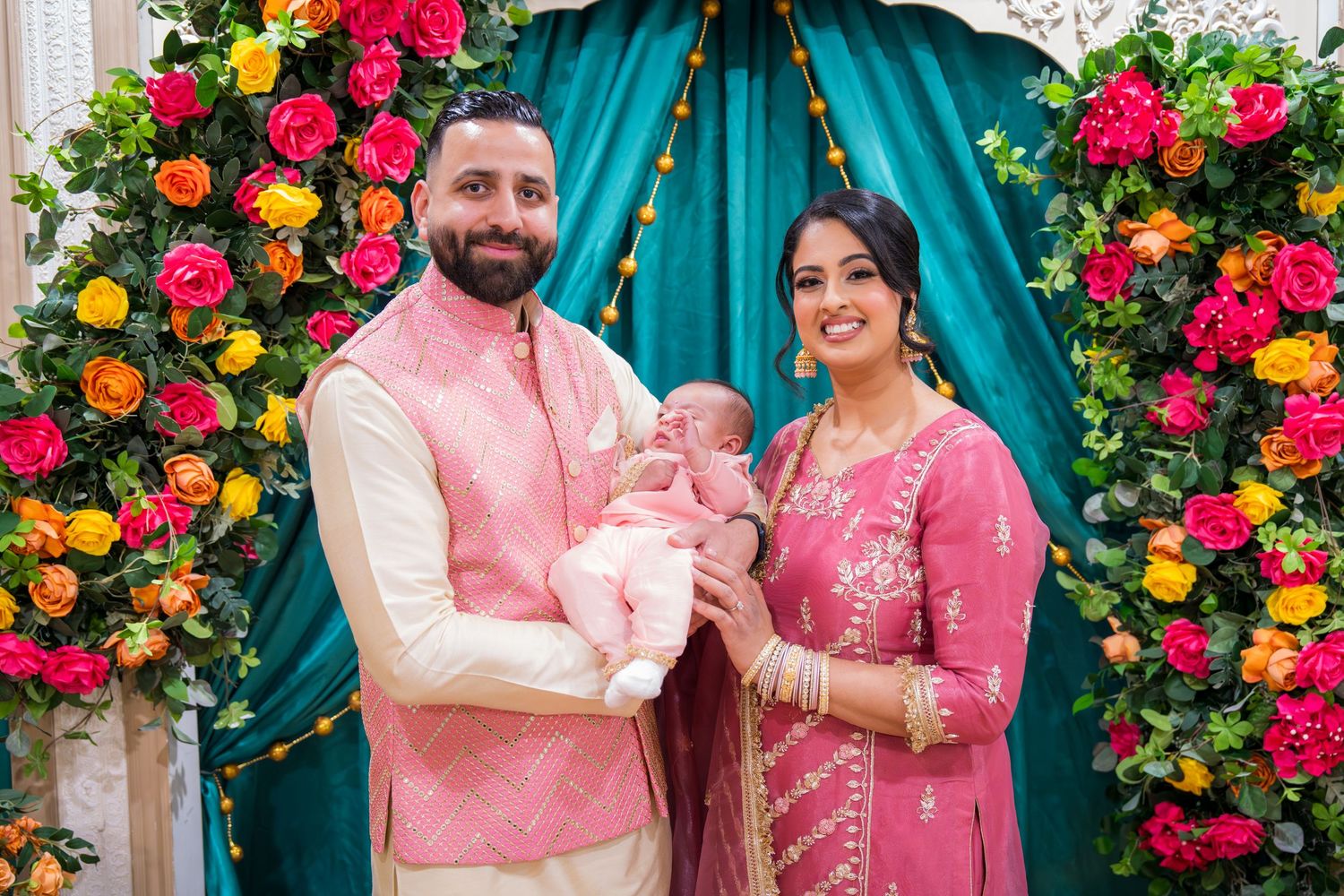 A couple in pink Indian attire holds their newborn baby in front of a floral arch with teal draping.