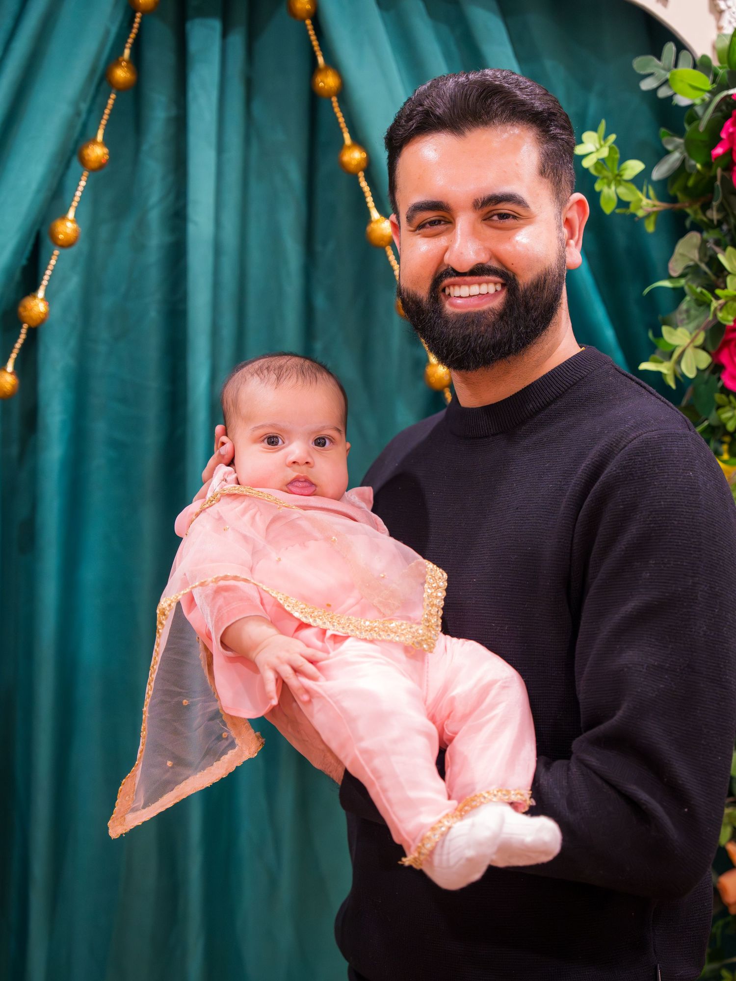 Smiling bearded man holding adorable baby in pink traditional outfit at South Asian celebration.