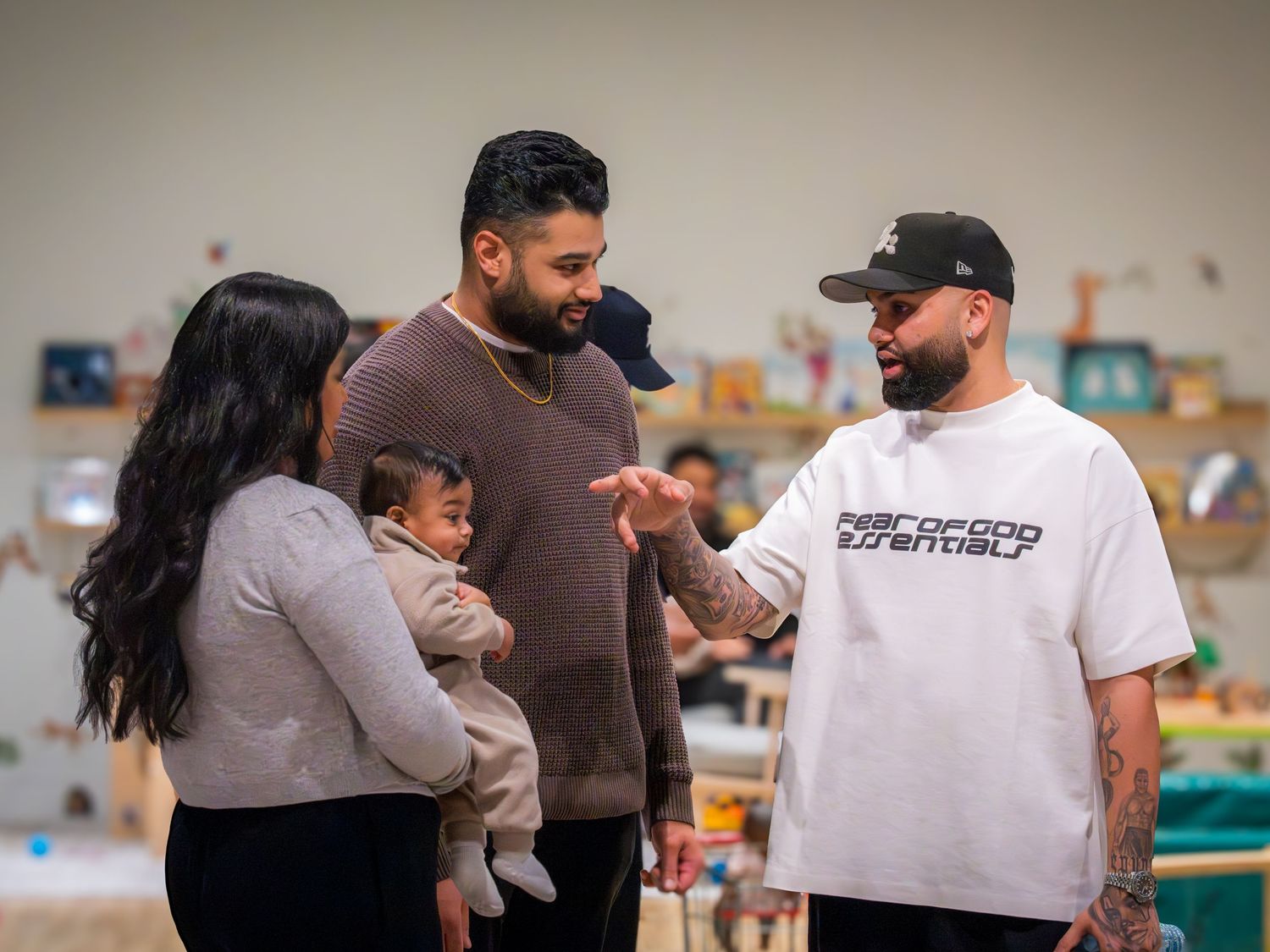 A man in a white graphic tee speaks with a couple holding a baby in a bright indoor space.