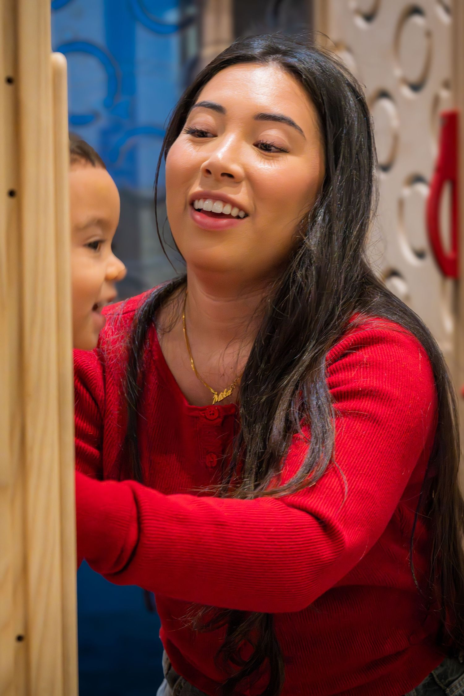 Smiling young woman with long black hair in red sweater interacting with a child at an indoor play area.