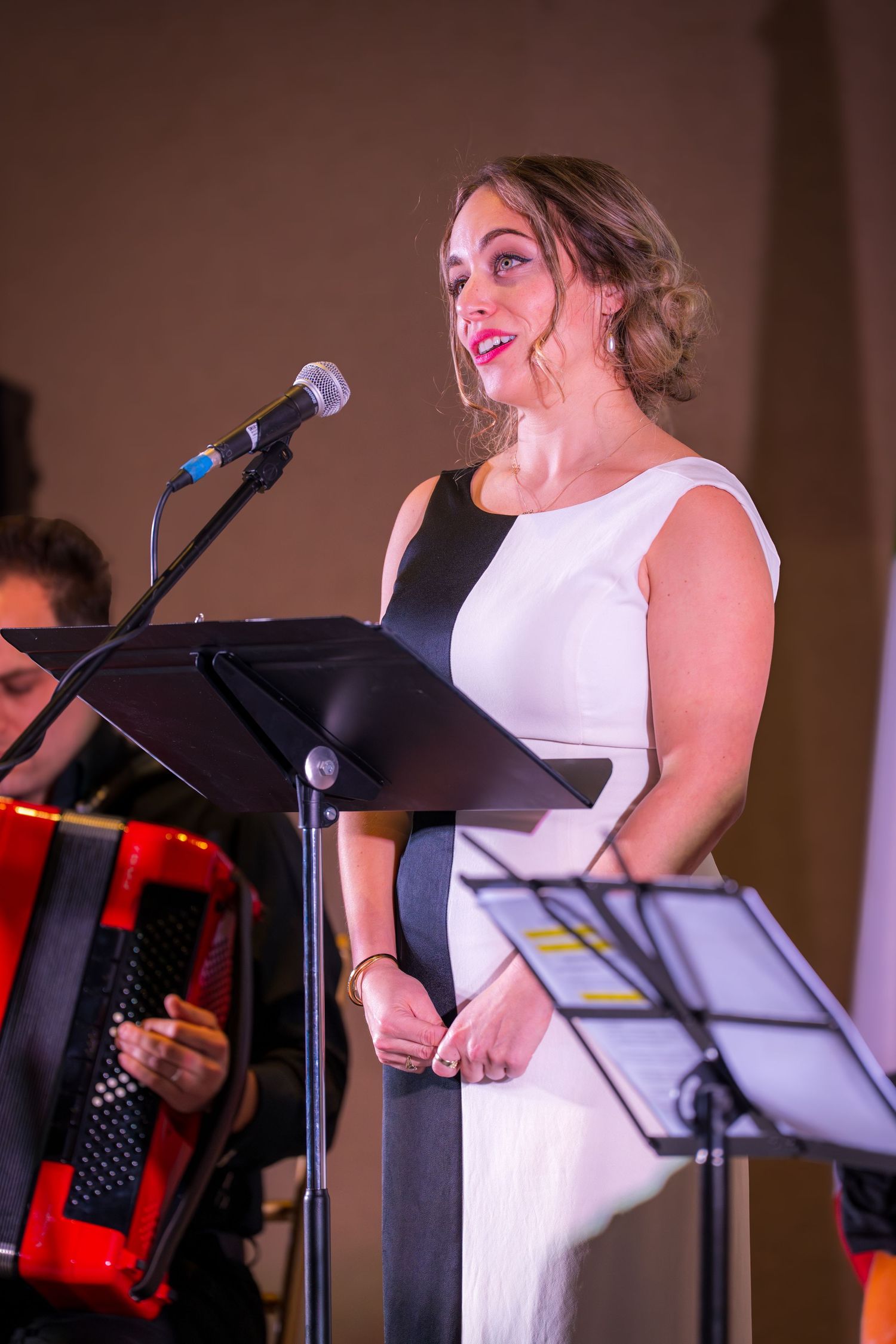 A woman in a black and white dress sings at a microphone stand during a live musical performance on stage.