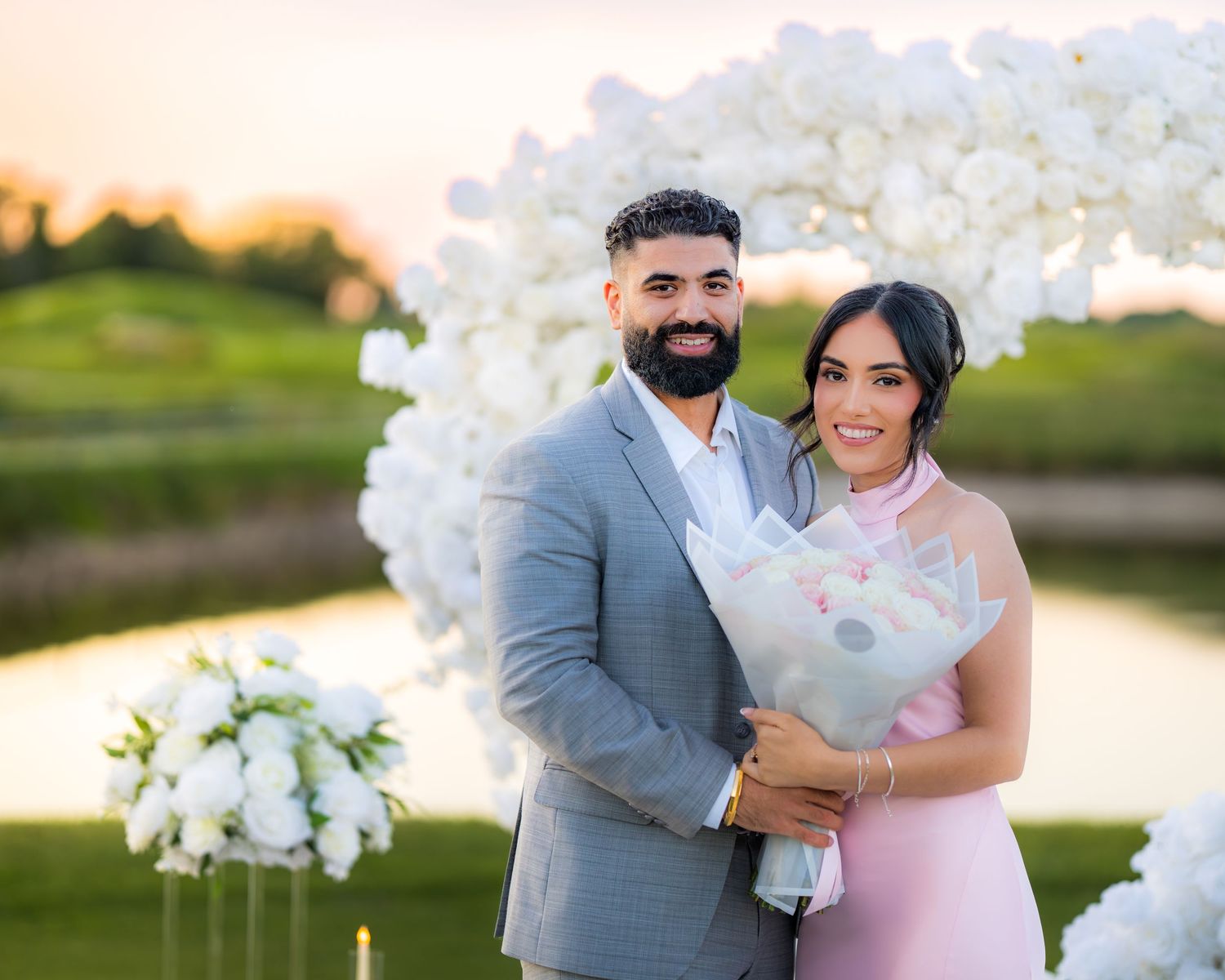 Joyful engagement celebration under white floral arch with sunset lighting.