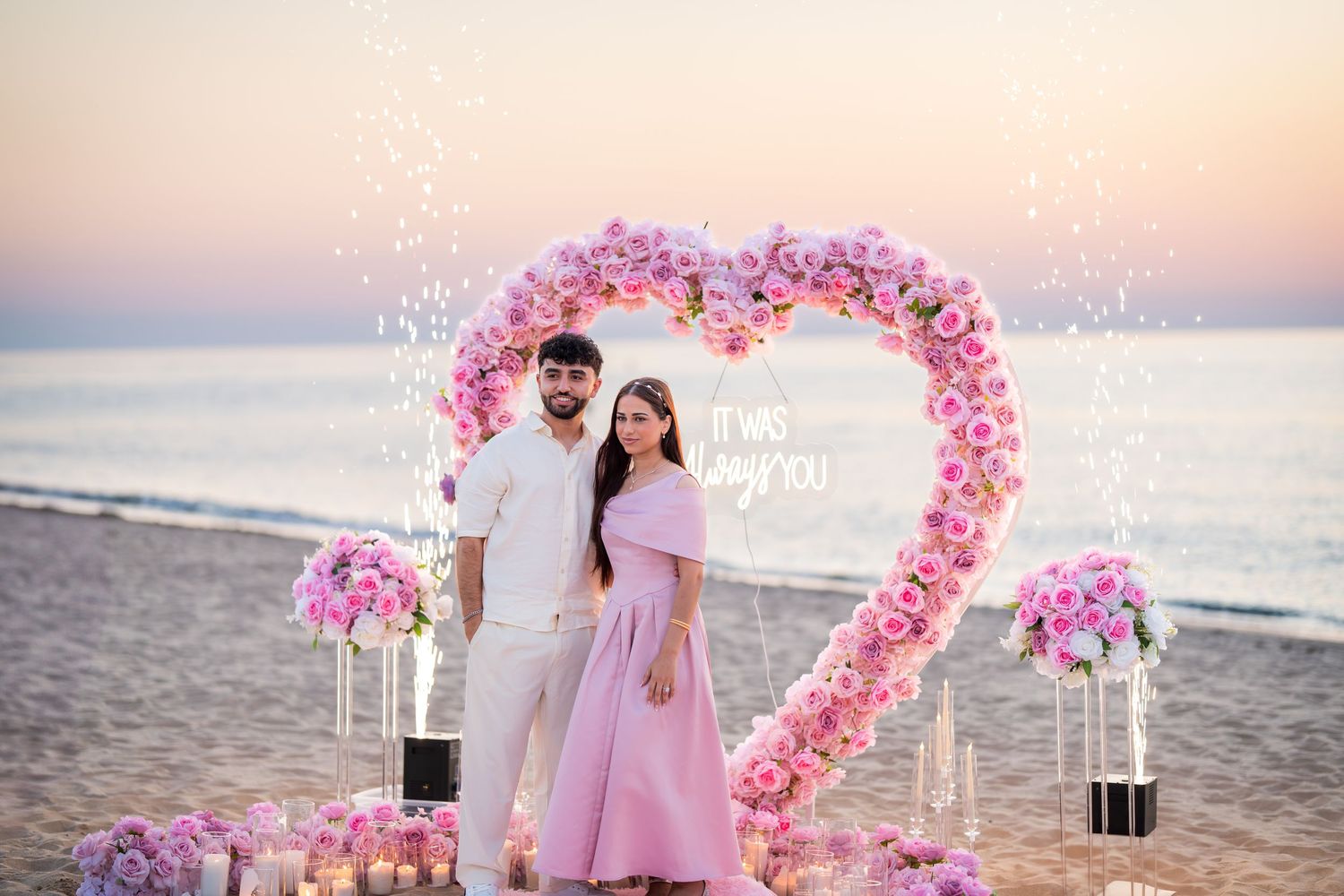 Elegant beach proposal setup featuring a heart-shaped floral arch and glowing candles at sunset.