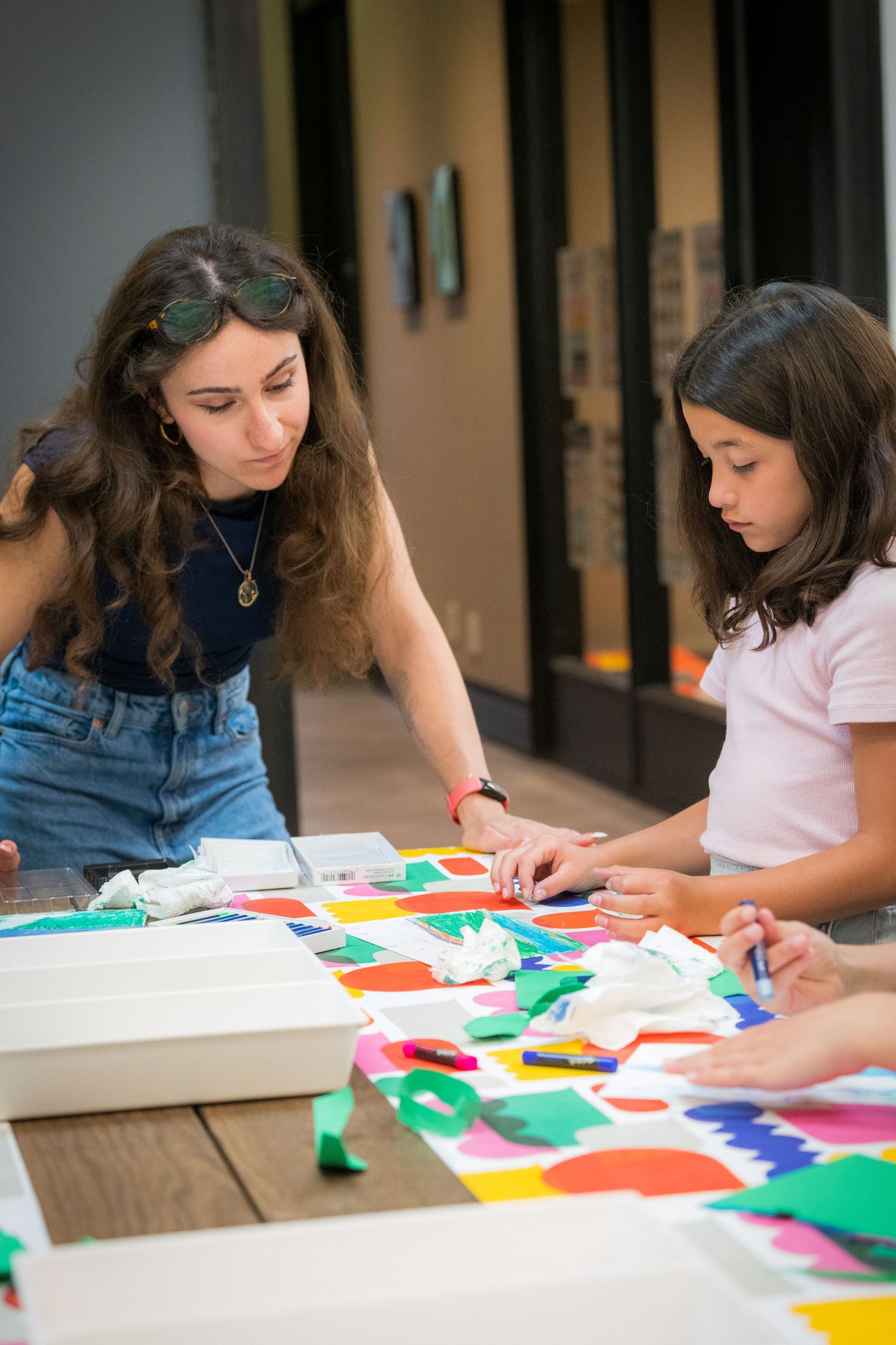 A woman instructor guides a young girl in creating a colorful collage art project at a table.