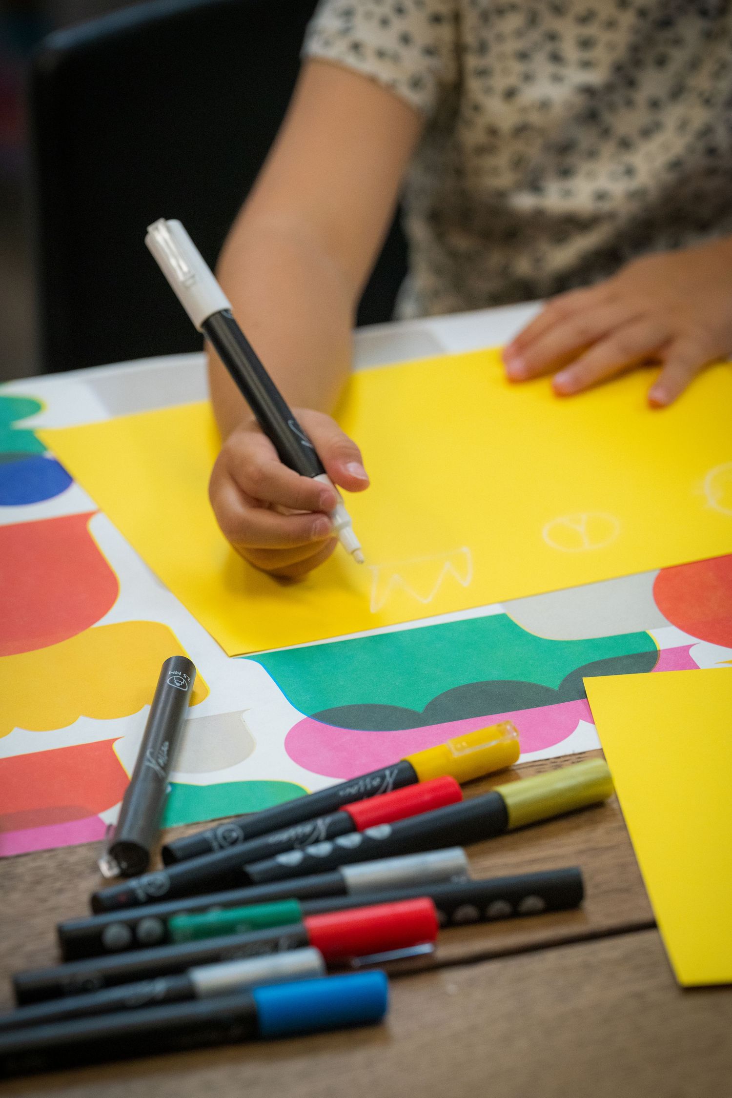 Child drawing on yellow paper with a white marker, surrounded by colorful markers on a table.