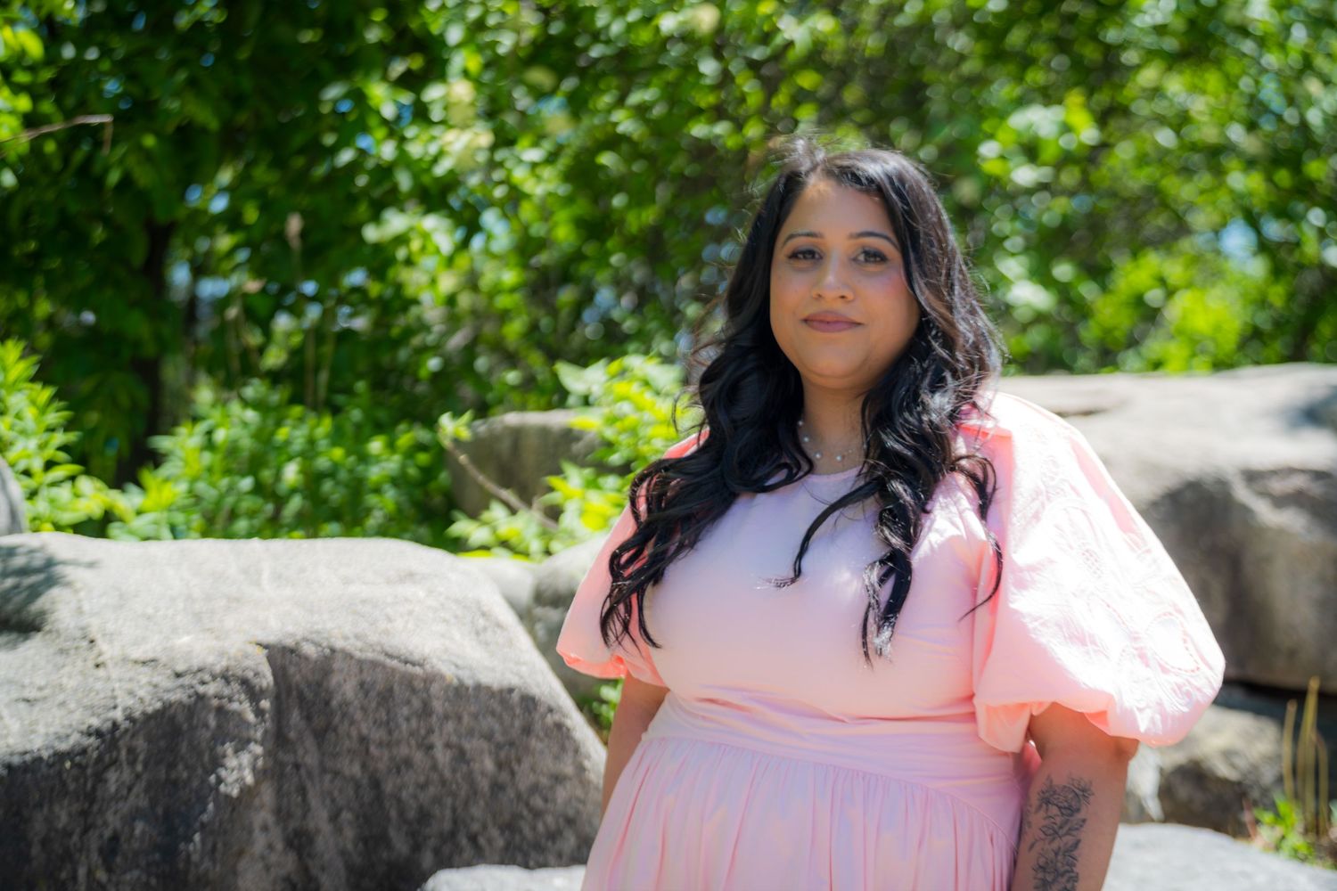 A woman in a pink puff-sleeve dress poses outdoors among green trees and large rocks.