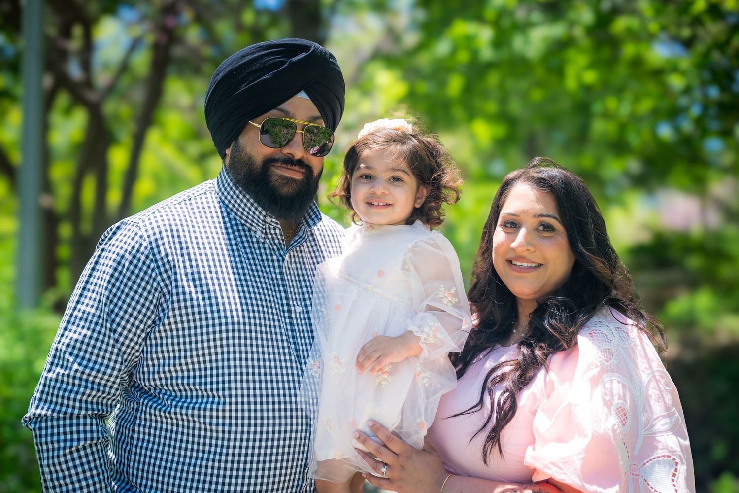Sikh family with man in turban, woman in pink, and young daughter in white dress outdoors.