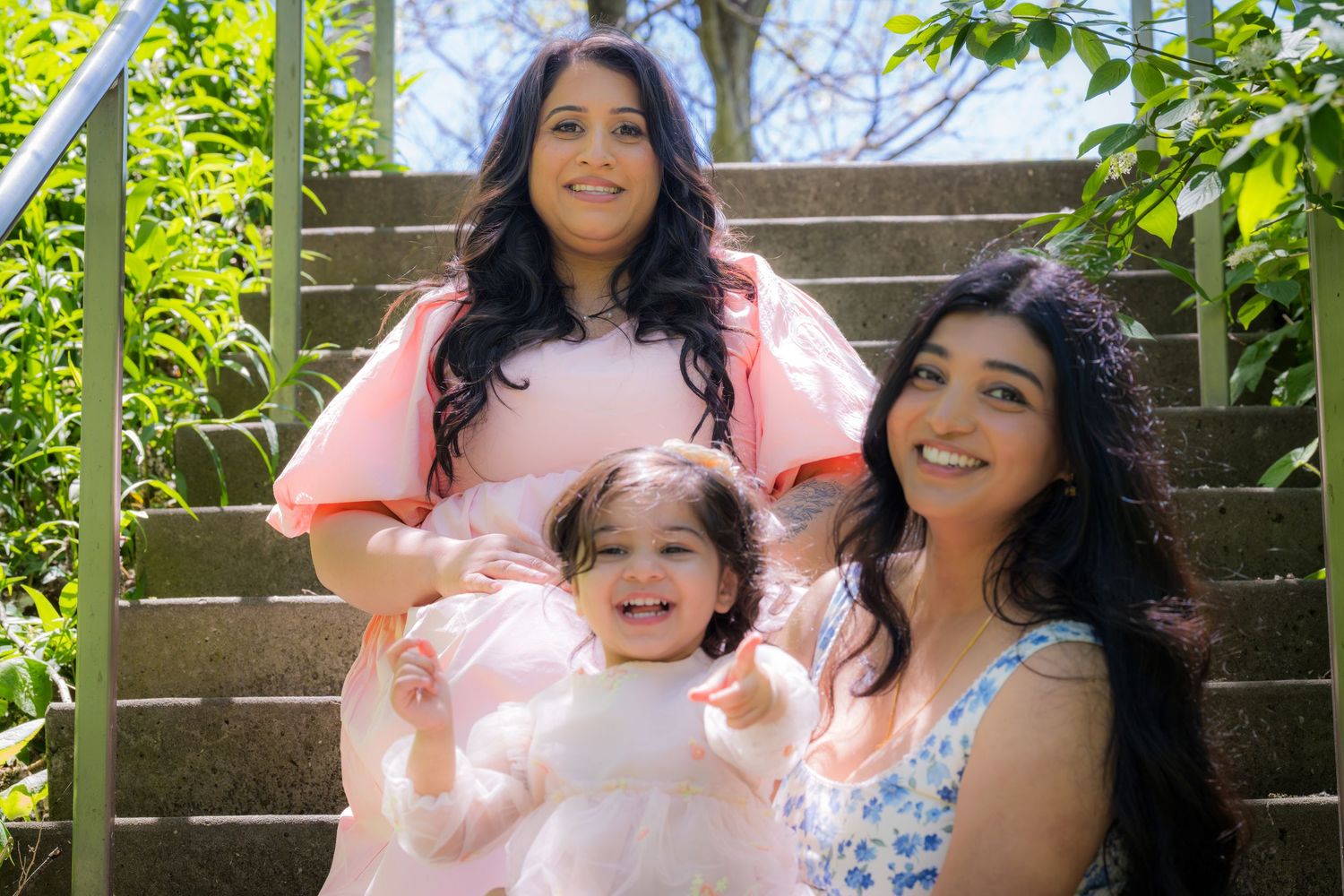 Two women in dresses sit on outdoor steps with a smiling toddler between them.