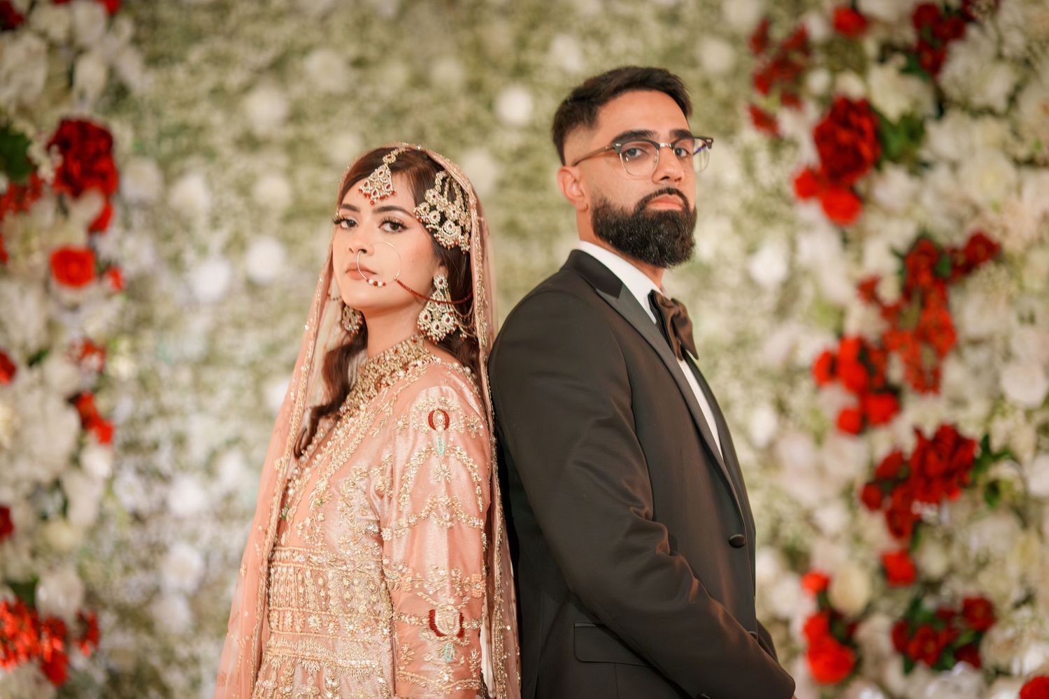 Stylish wedding couple poses against white and red floral backdrop in traditional attire.