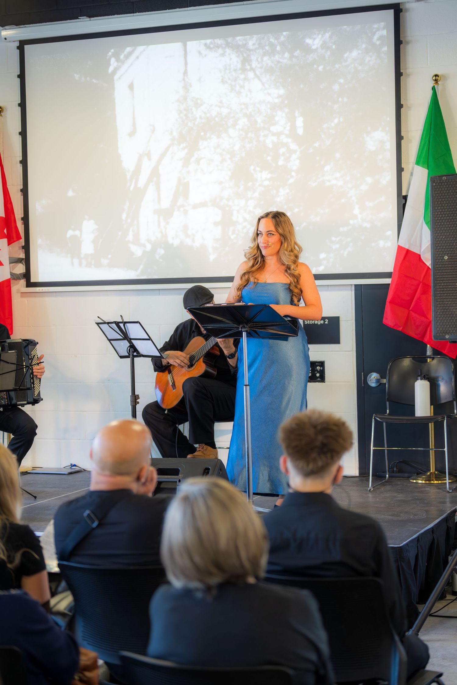 Elegant woman in blue dress speaking at podium with guitarist and Italian flags at formal event.