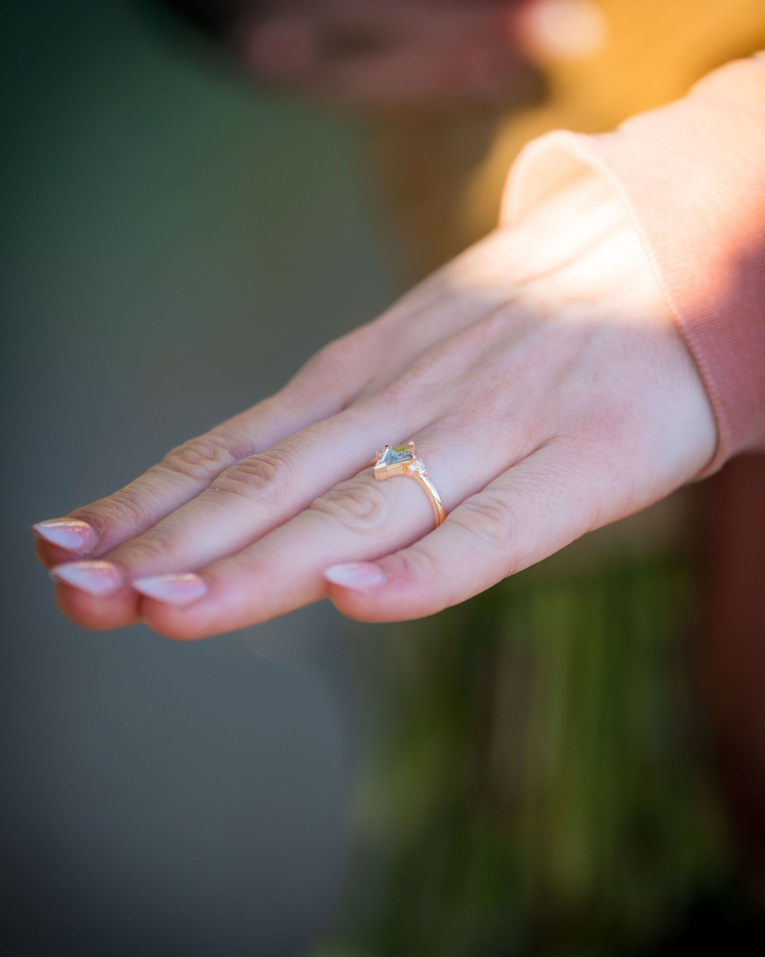 Close-up of a woman's hand displaying a delicate gold solitaire engagement ring outdoors.