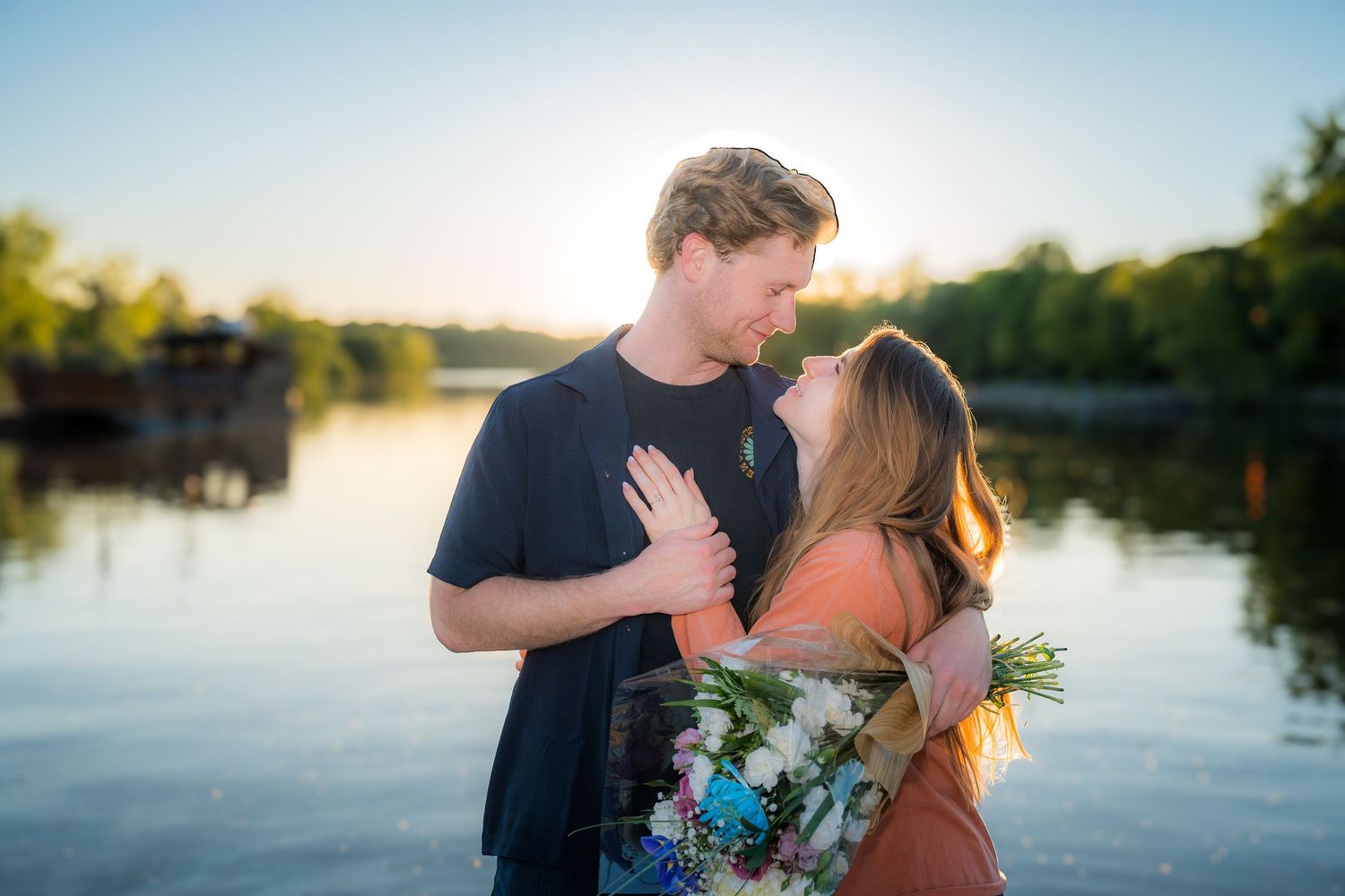 Couple sharing a romantic almost-kiss at sunset by a serene lake, woman holding a colorful bouquet.