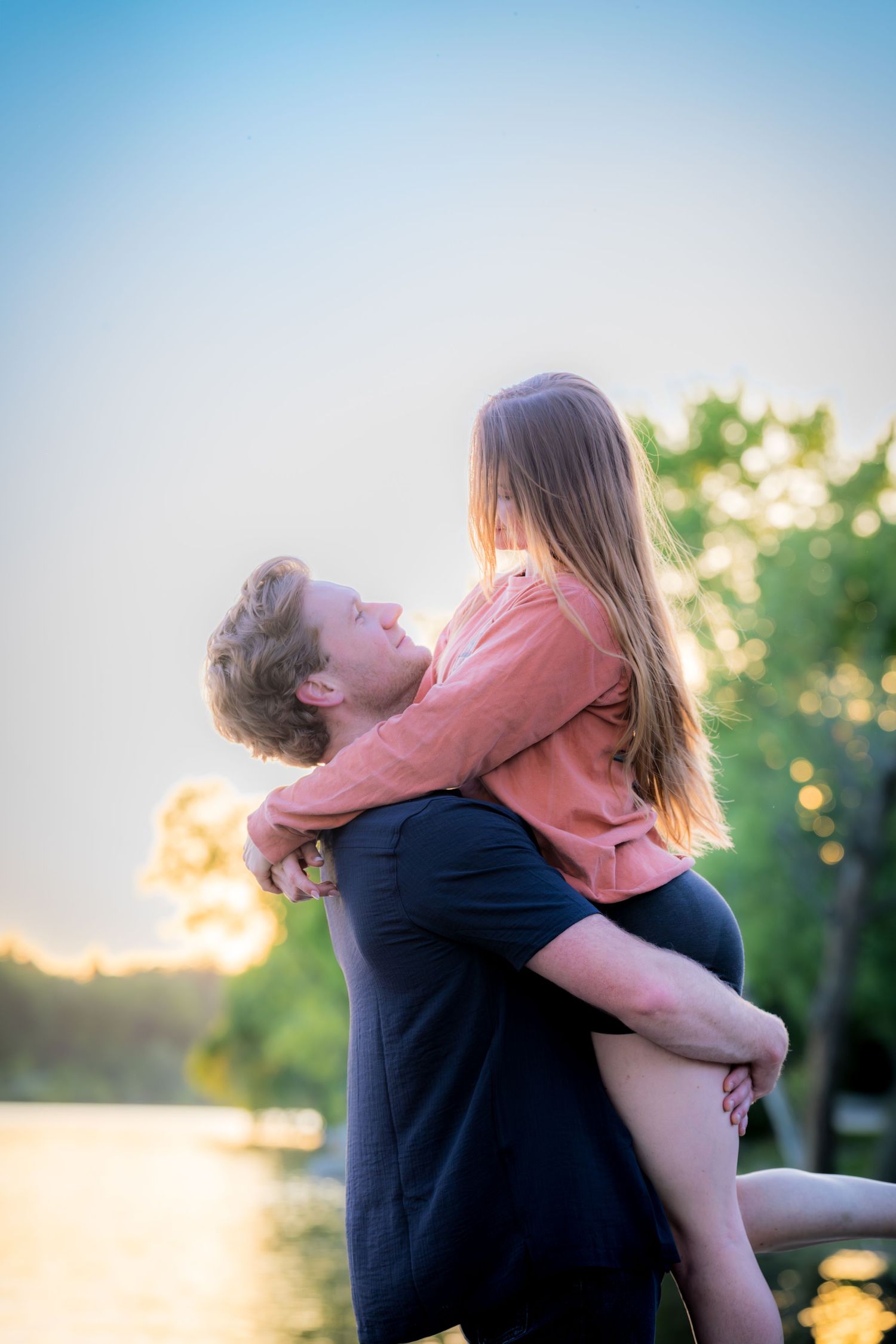 Joyful couple embracing outdoors by a lake surrounded by lush trees at golden hour.
