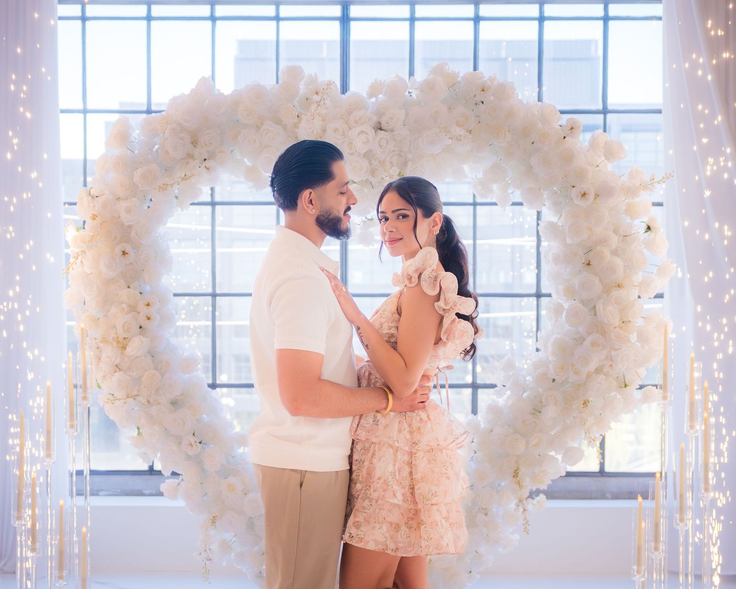 Couple embracing in front of a white floral heart arch with fairy lights at a romantic engagement photo shoot.