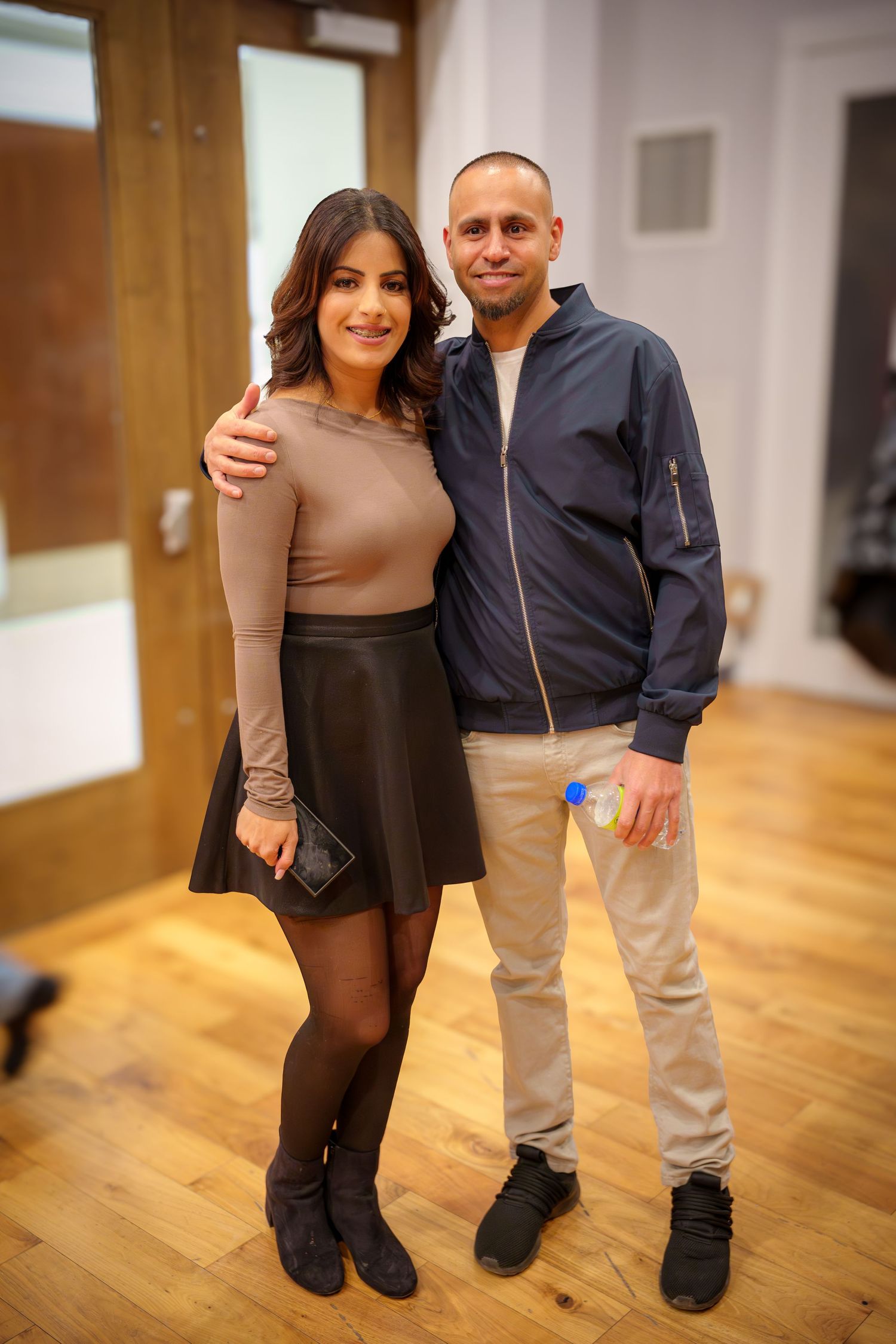 Two people pose together on a hardwood floor in casual attire showing height difference.