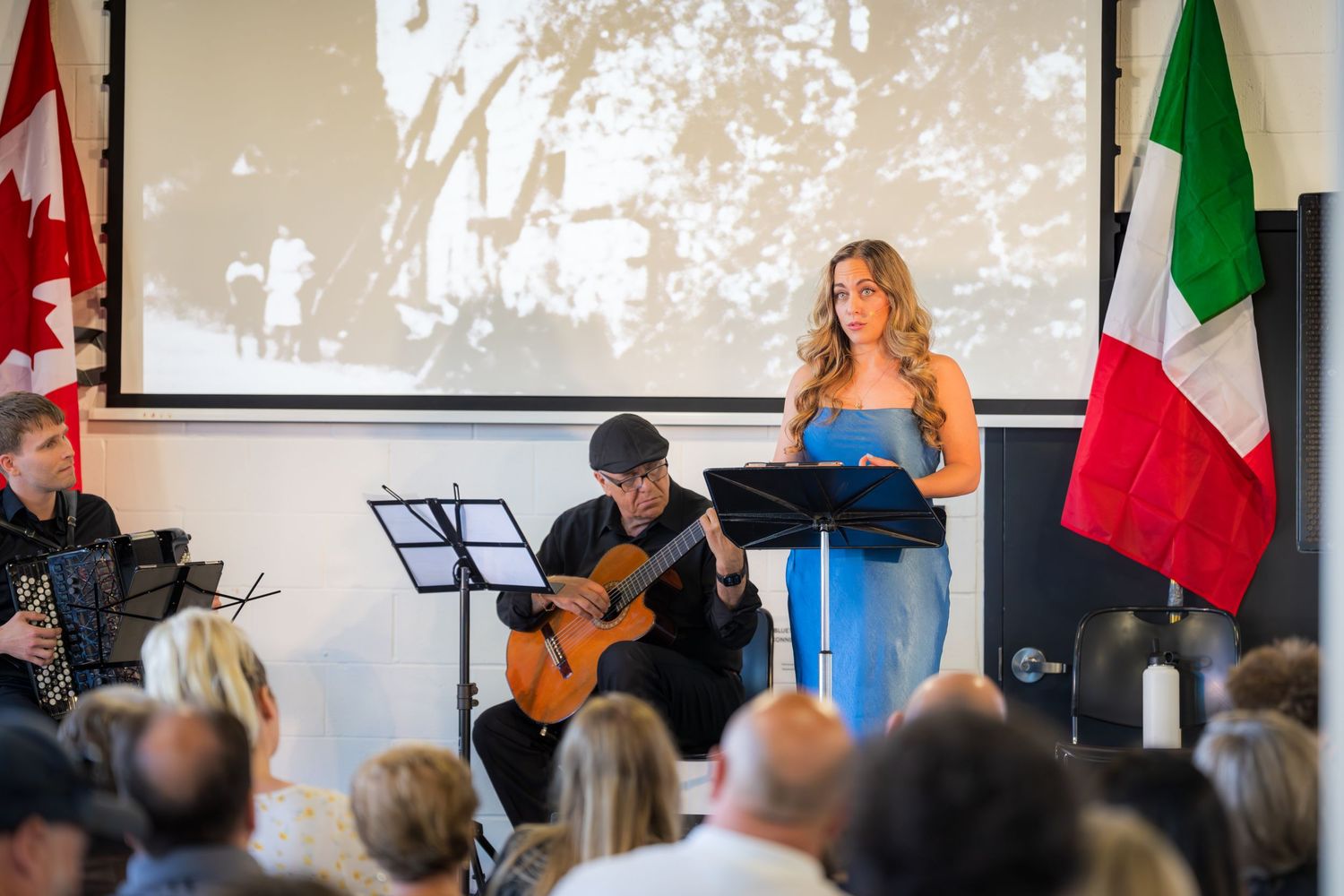 Woman singing at podium with guitarist at Italian-Canadian cultural event, Canadian and Italian flags displayed.