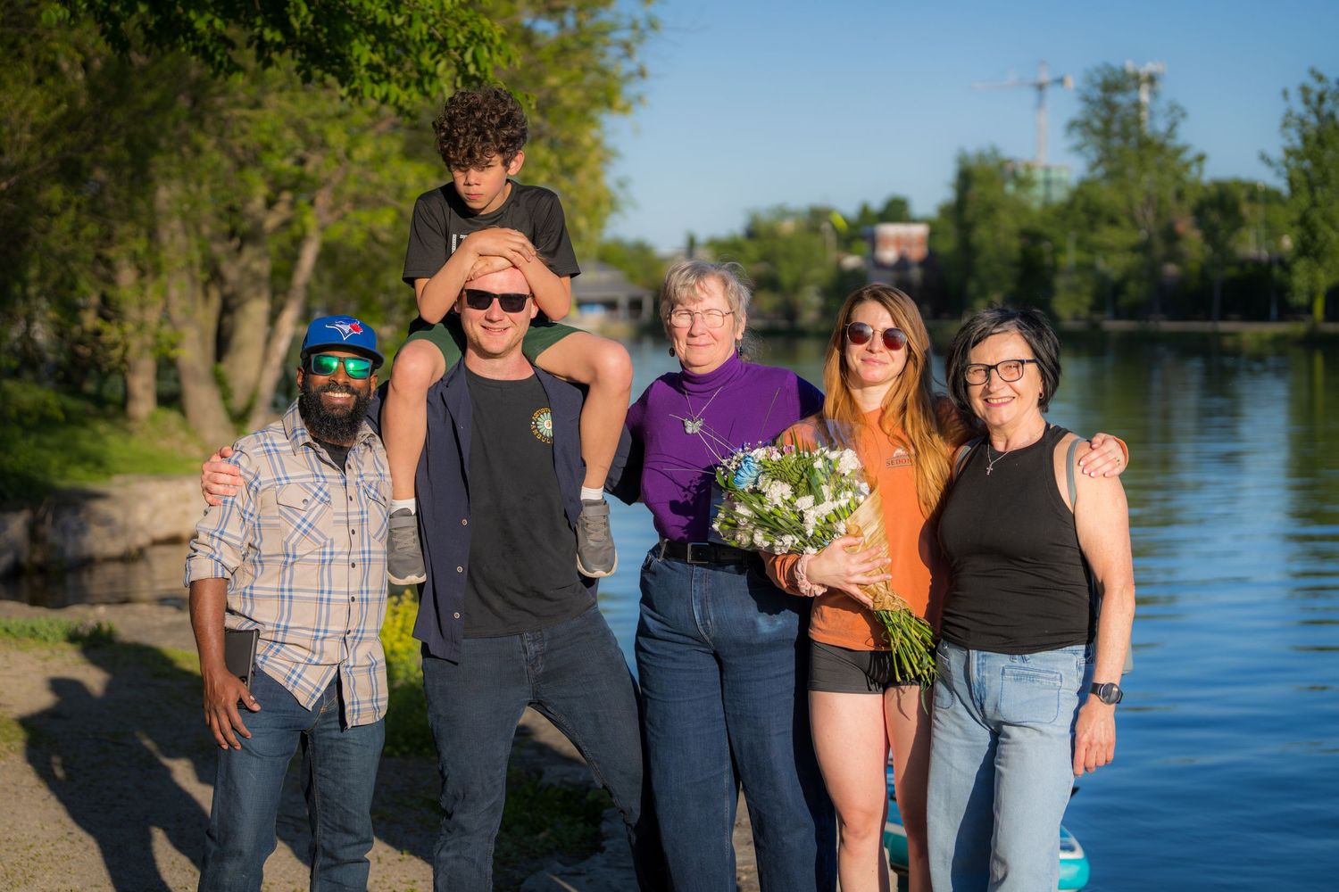 Group of six people posing together outdoors near a river on a sunny day, one holding a bouquet.