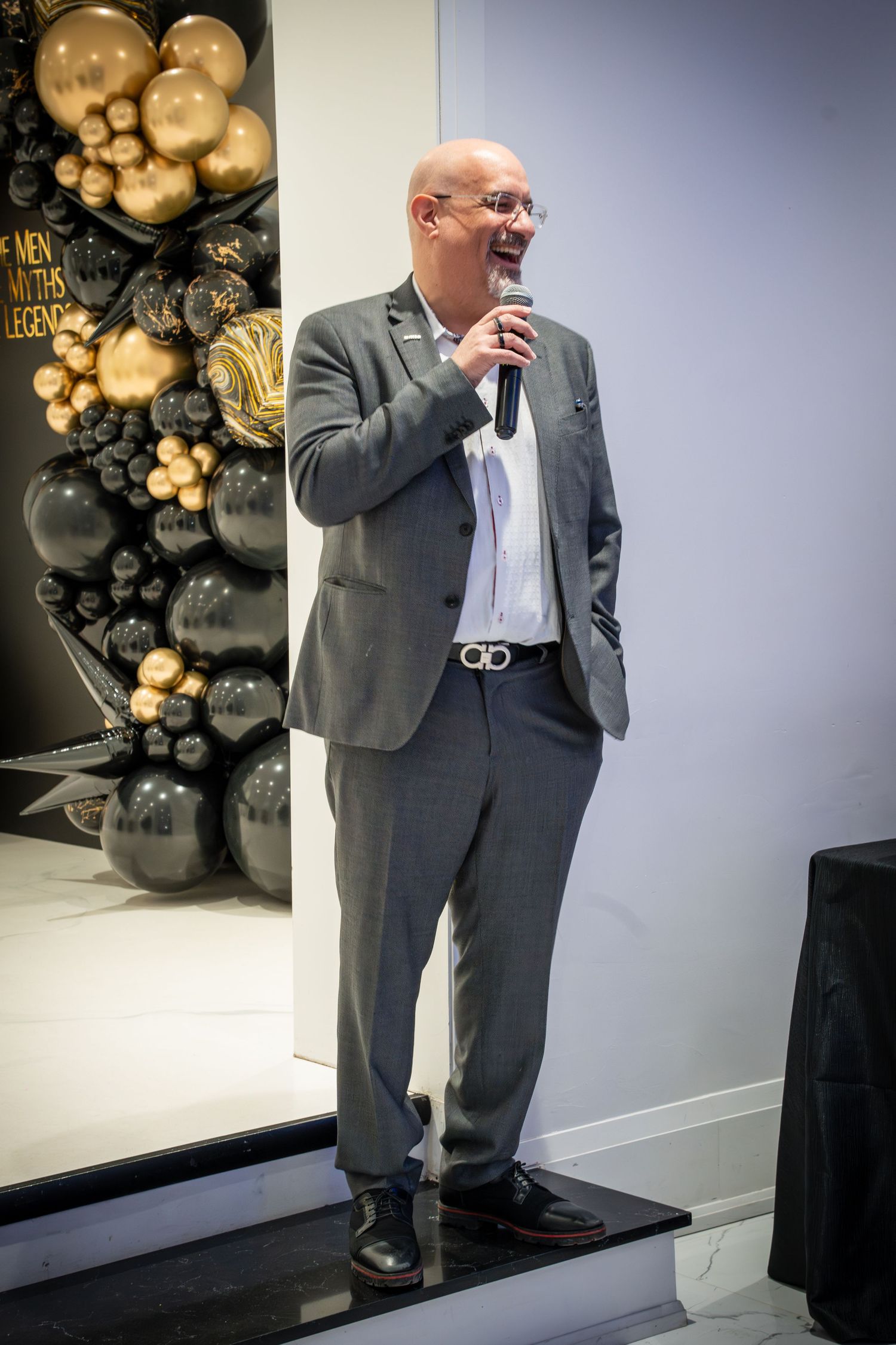 A presenter in a grey suit speaks from a podium with decorative black and gold balloons in background.