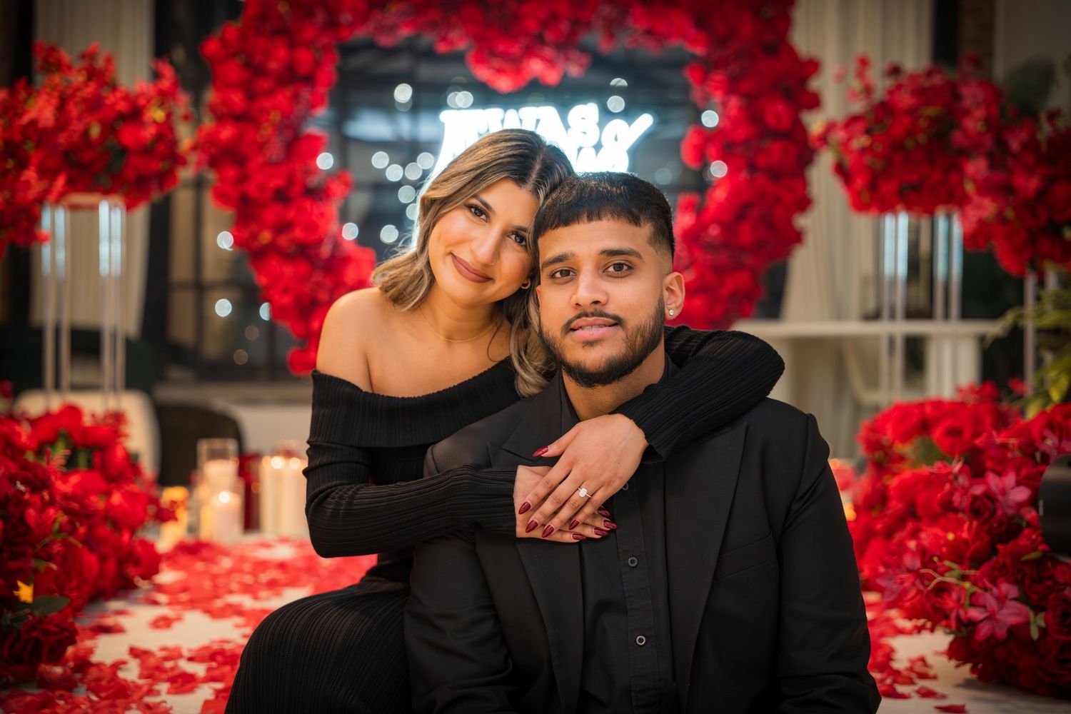Couple poses intimately in front of red rose wall with neon lighting and romantic ambiance.