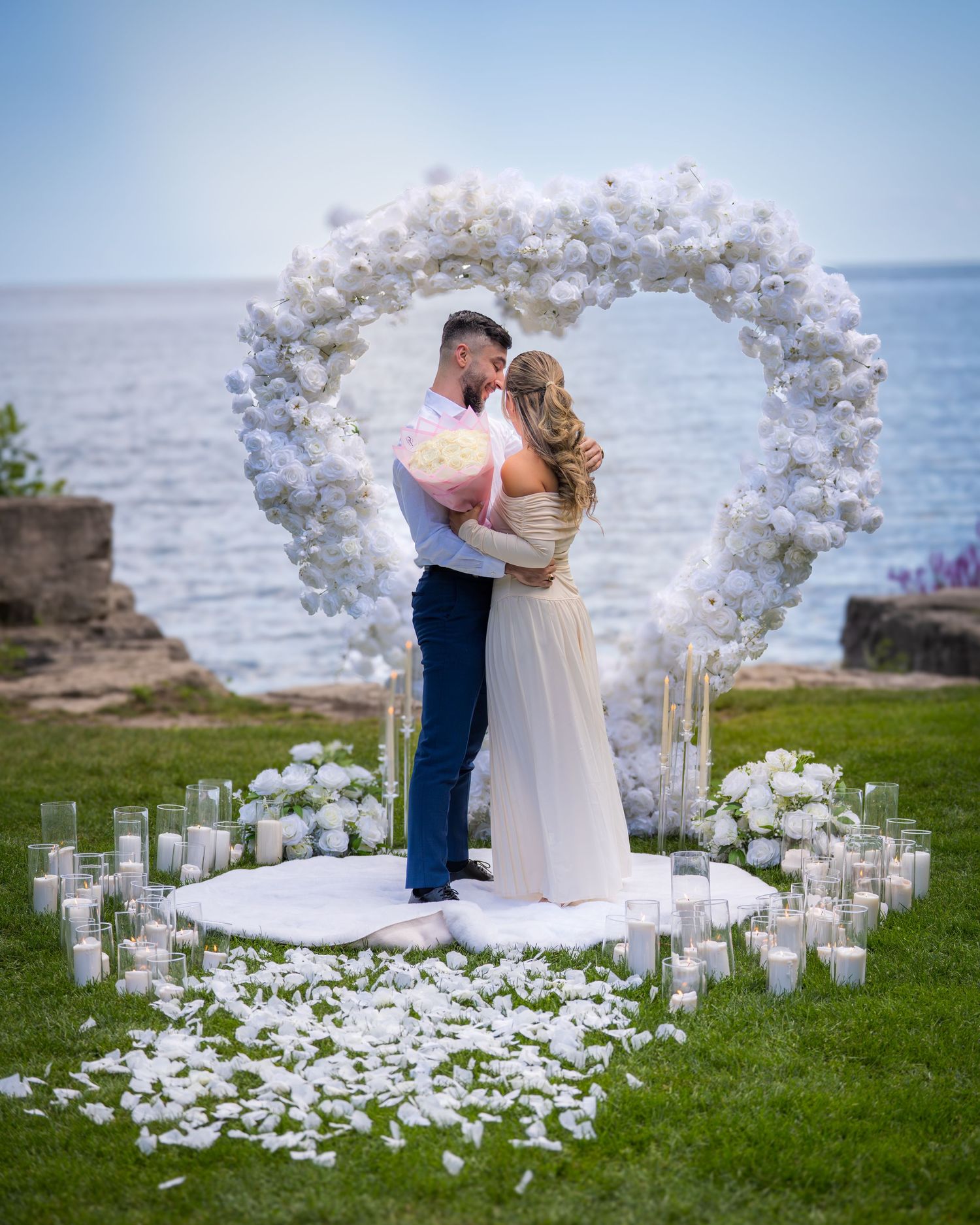 Coastal wedding setup featuring white flower arch and pathway lined with candles against ocean backdrop.