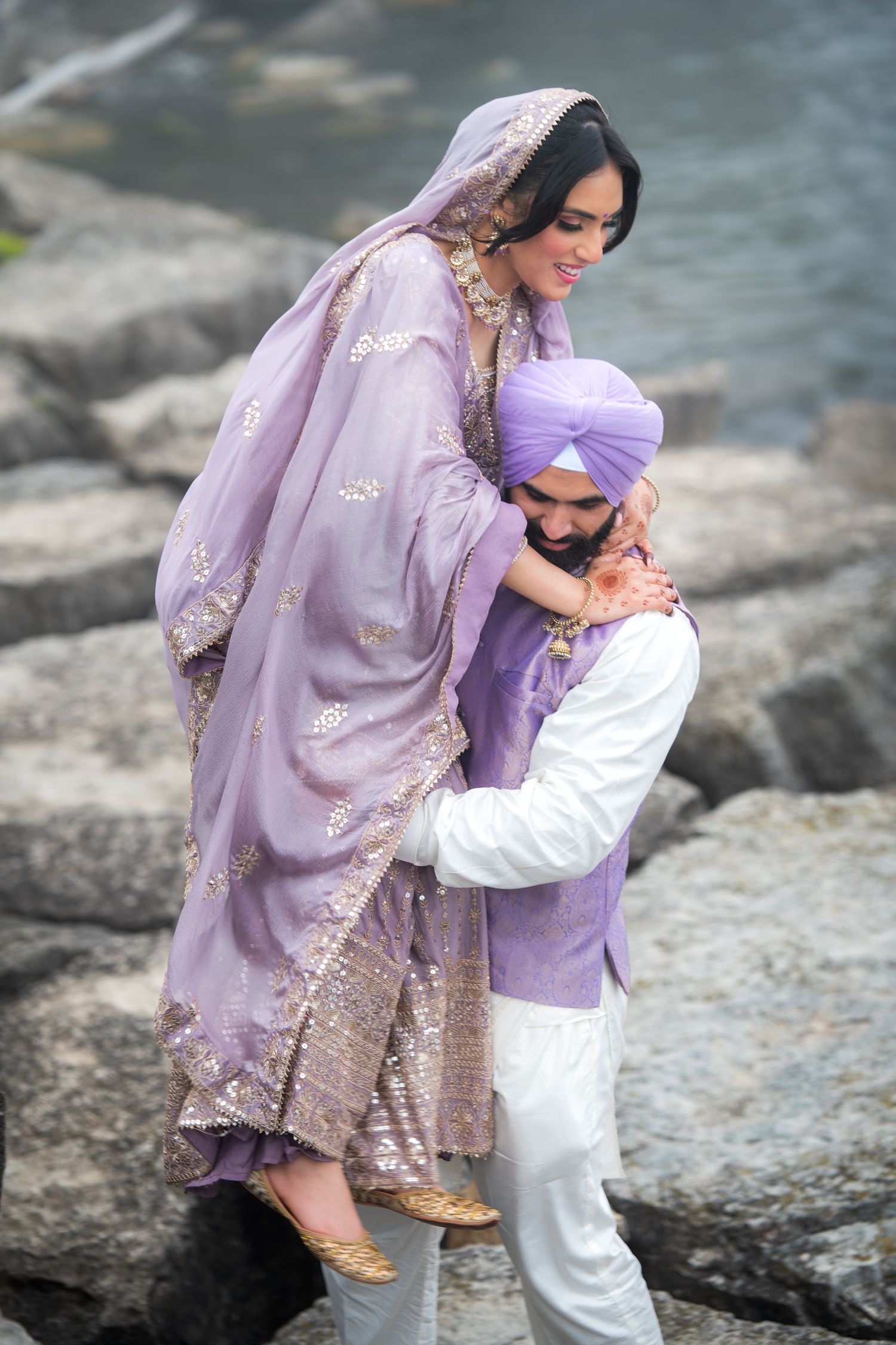 Romantic couple in traditional purple attire embrace on rocky shoreline.