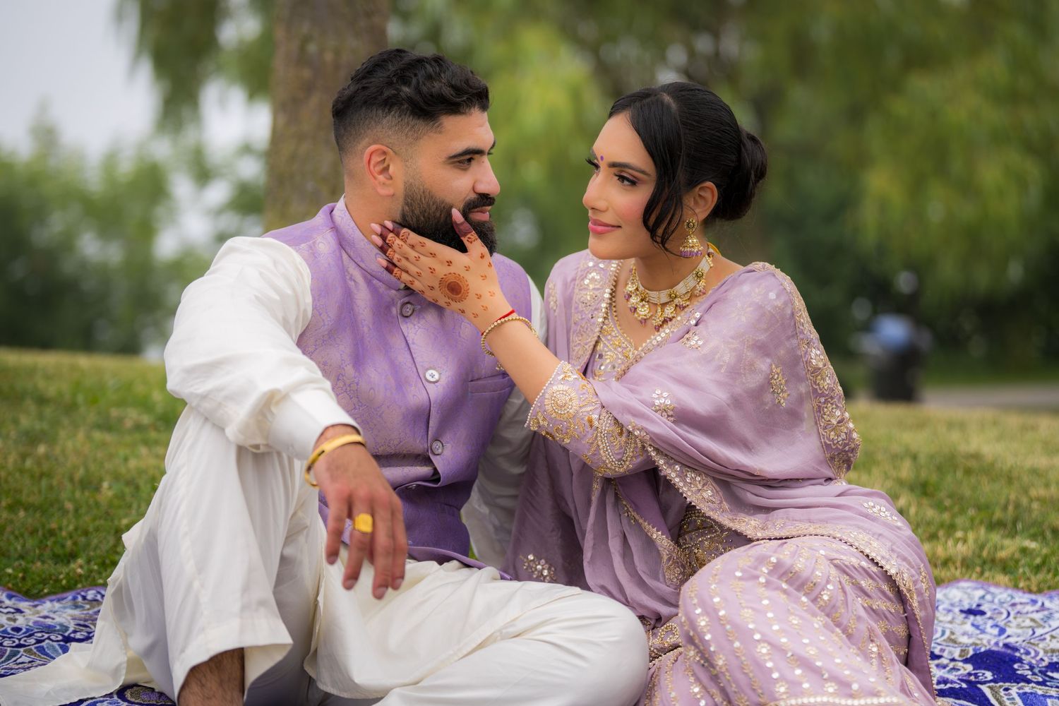 Romantic couple in coordinated purple and white attire pose on decorative blanket in park setting.