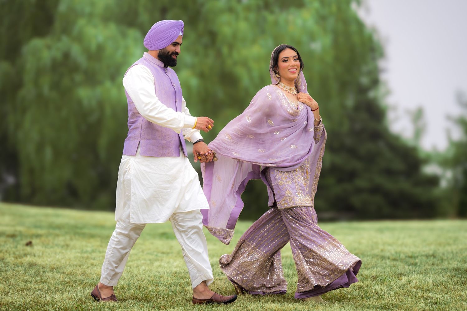 Two people in elegant purple and white traditional outfits dance together on a lush green hillside.