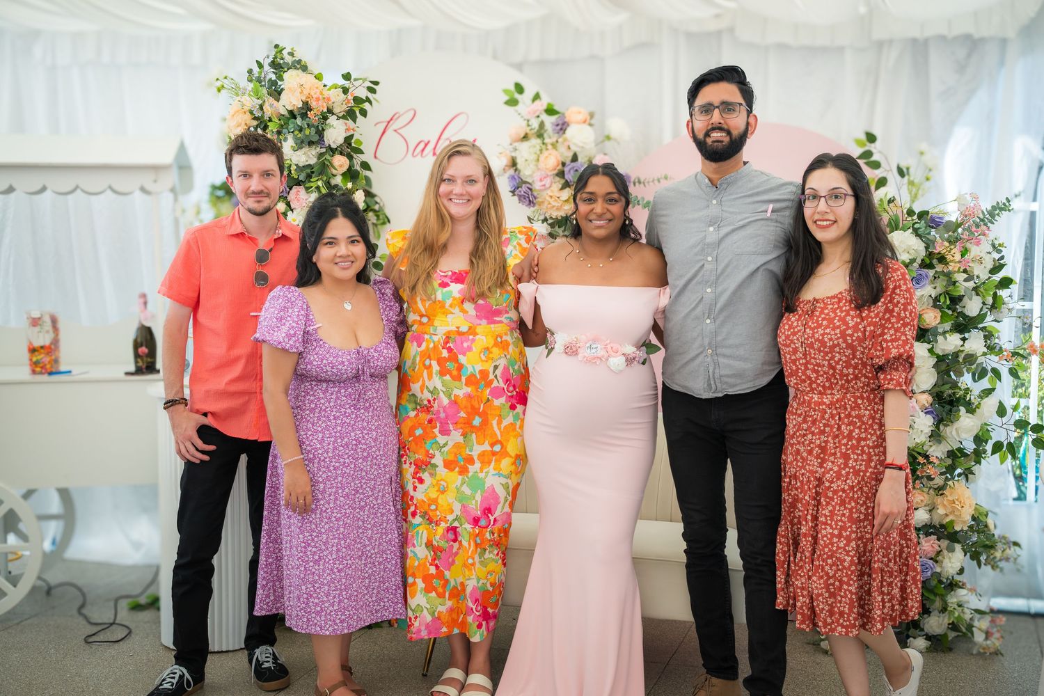 Group photo at a baby shower with floral arch backdrop and people wearing colorful spring dresses.