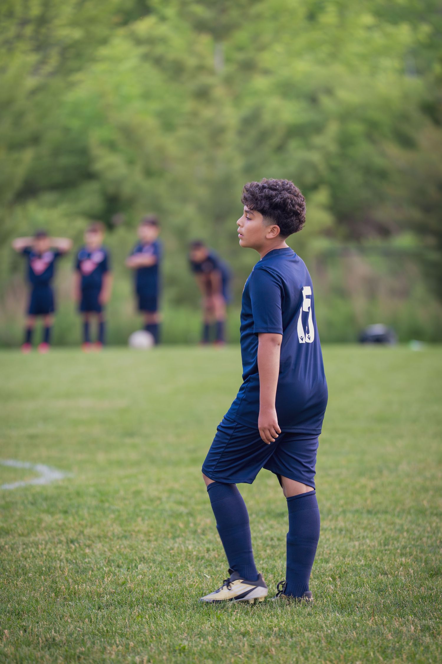 A player in navy blue uniform performs warmup exercises on a soccer field with teammates in background.