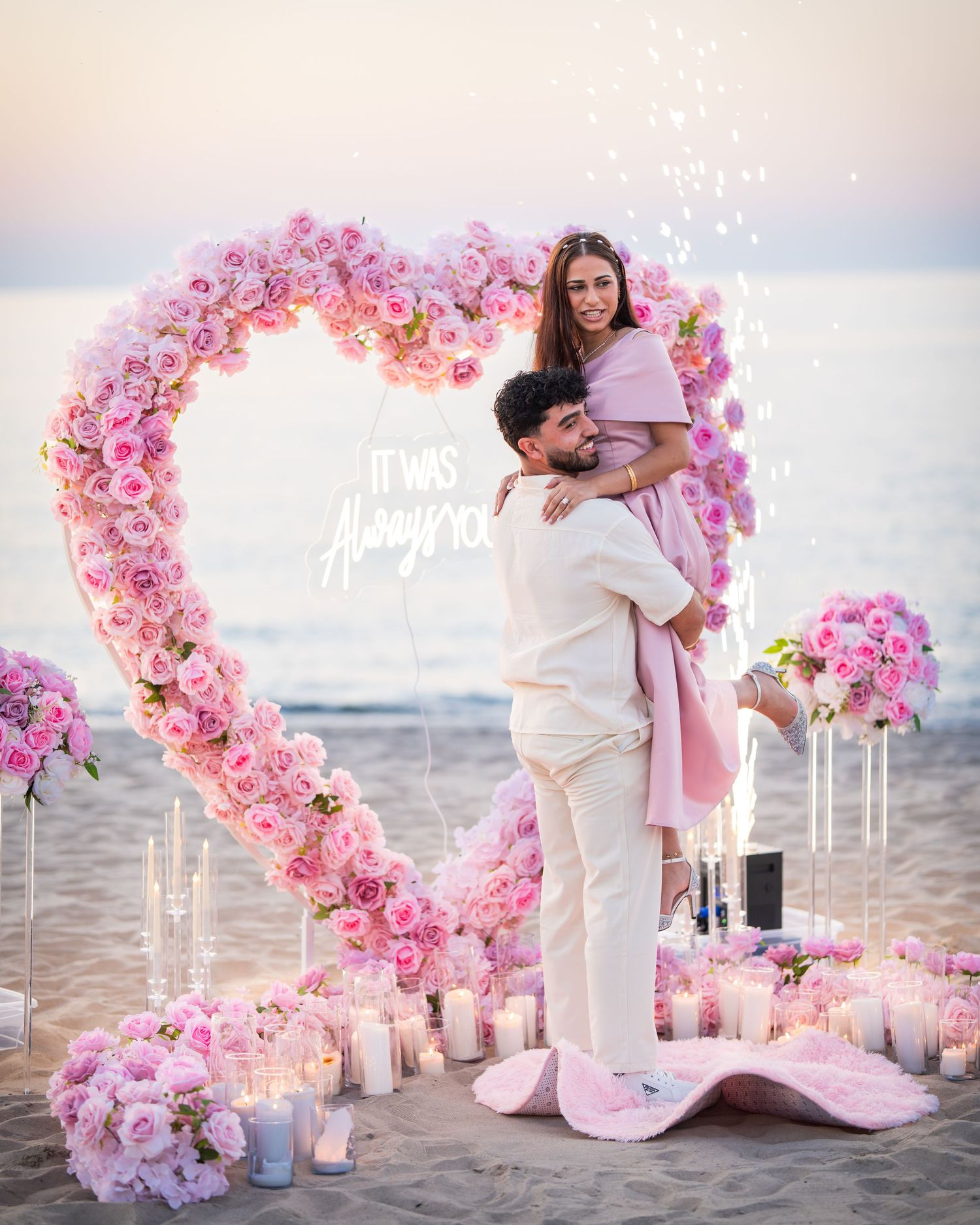 A romantic beach moment captured through a pink floral heart arch with candles and sunset lighting.